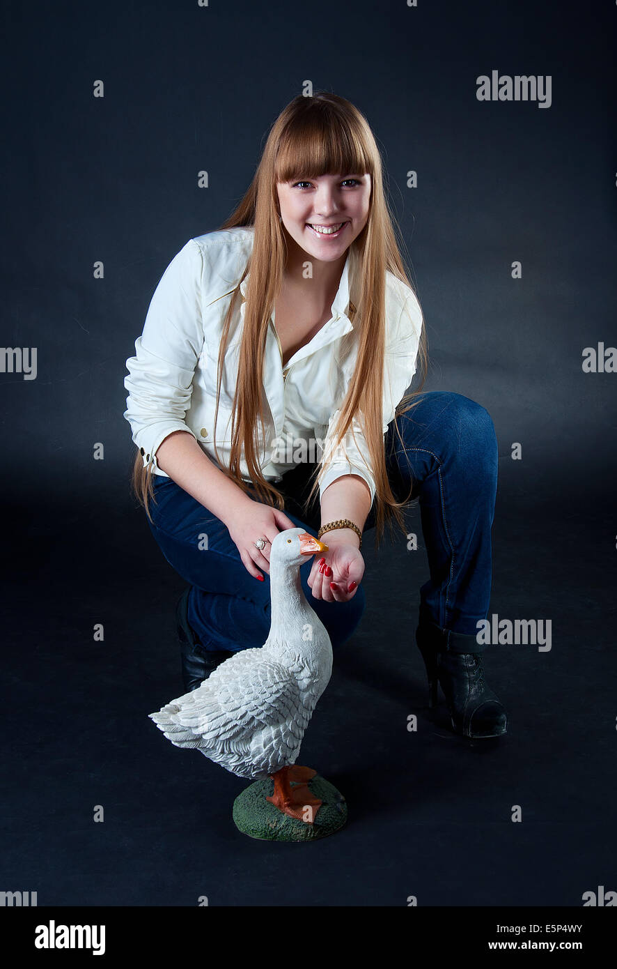 Beautiful caucasian girl with long blond hair wearing white jacket and blue jeans and feedenig the artificial goose Stock Photo