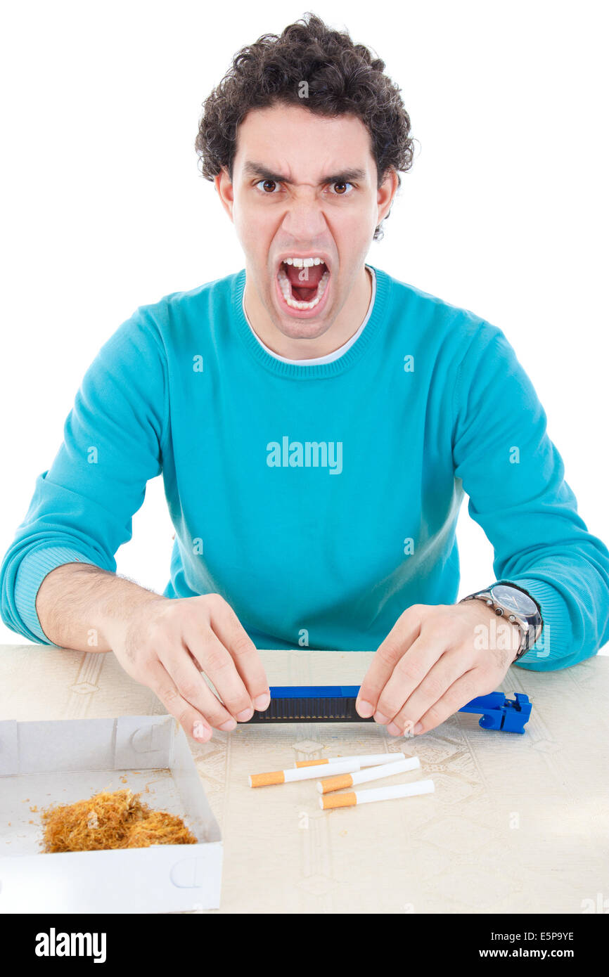 Angry man in blue sweater making cigarettes with device for cigar and dry tobacco on table, economic crisis due high price of ci Stock Photo