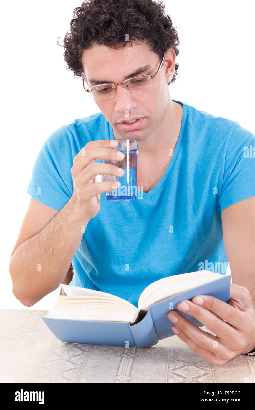 casual young man in blue t-shirt with glasses sitting at table and reading book while drinking healthy water from a glass, stude Stock Photo