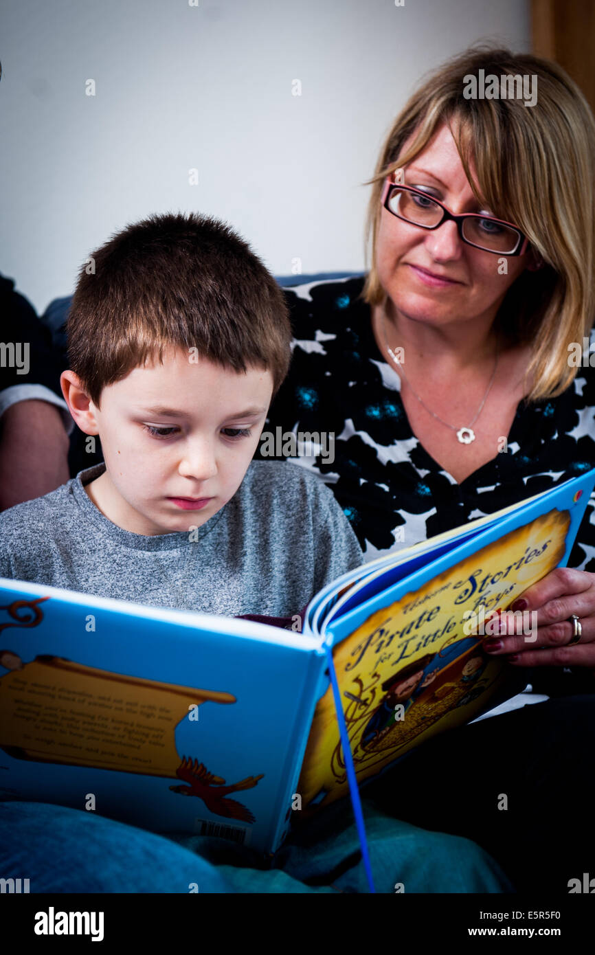 Boy 8 9 10 reading a book with mother listening in background Stock Photo
