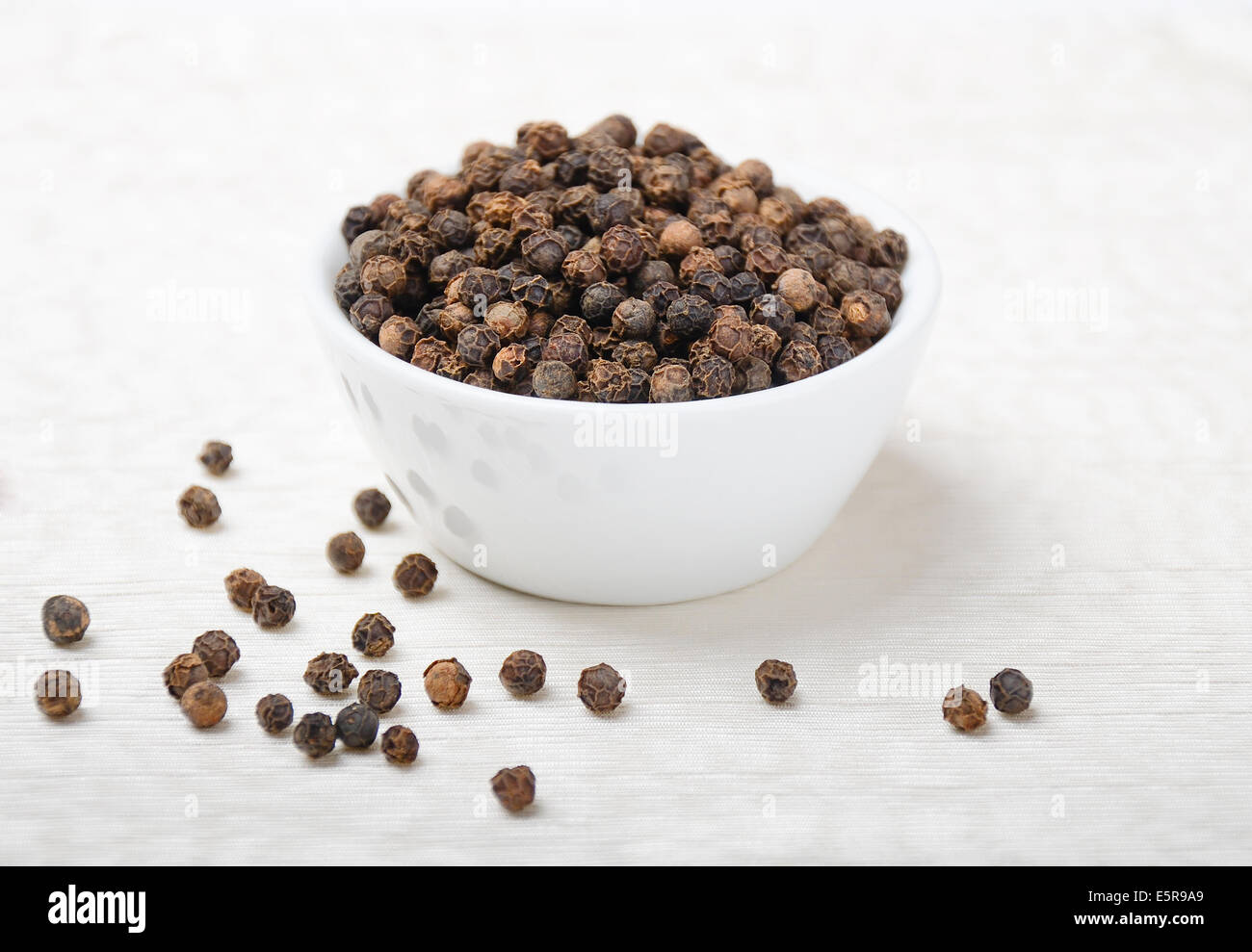 Black Pepper - Dried black peppercorns in a white porcelain bowl on white textile made of linen. Stock Photo