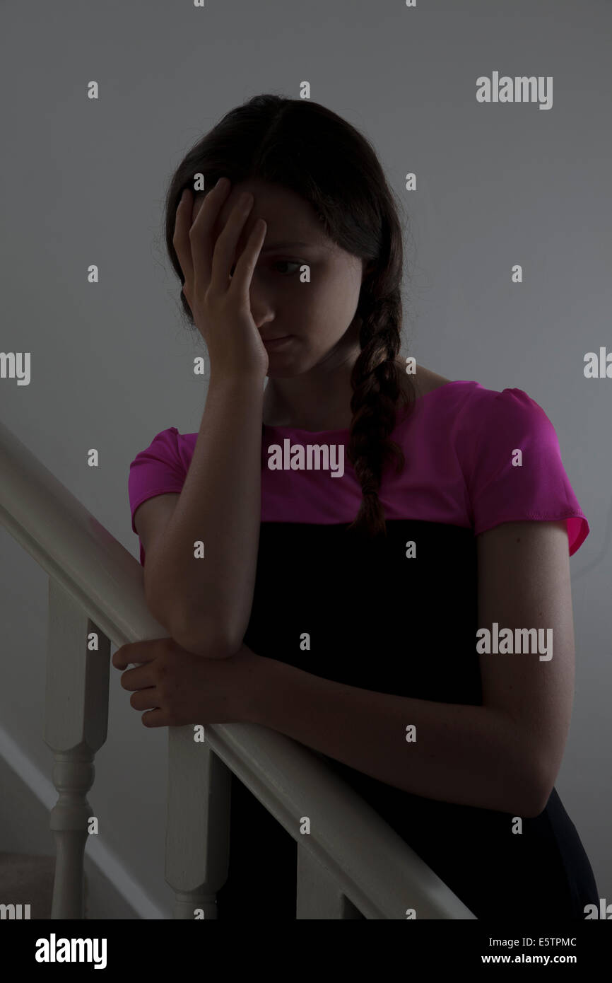 Young female wearing a pink top and hair in plats standing on the stairs indoors, hand on face. Stock Photo