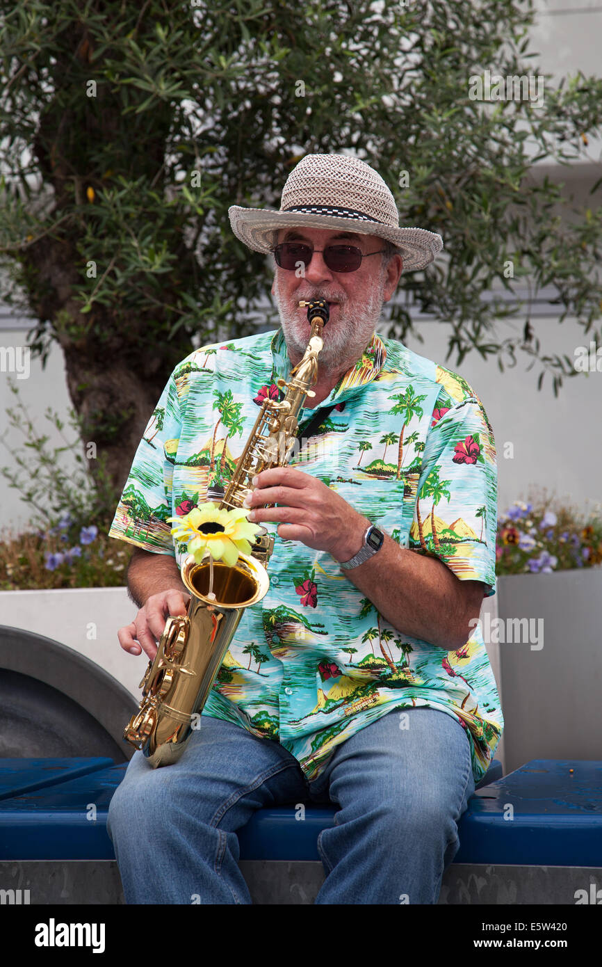 Manchester, UK 6th August 2014. 'Spud' Geffrey Seddon playing the Alto Saxophone at the Dig the City Deansgate, an annual urban gardening festival delivered by Manchester's Business Improvement District – BID.   Floral displays, bus stops and a Vimto garden transforming Manchester city centre as Dig the City returned.   Sixteen show gardens on display during the urban gardening festival, from August 2 to 10.  Each has been designed especially for the event, with their designers having to embrace the challenges of gardening in a small urban space. Stock Photo