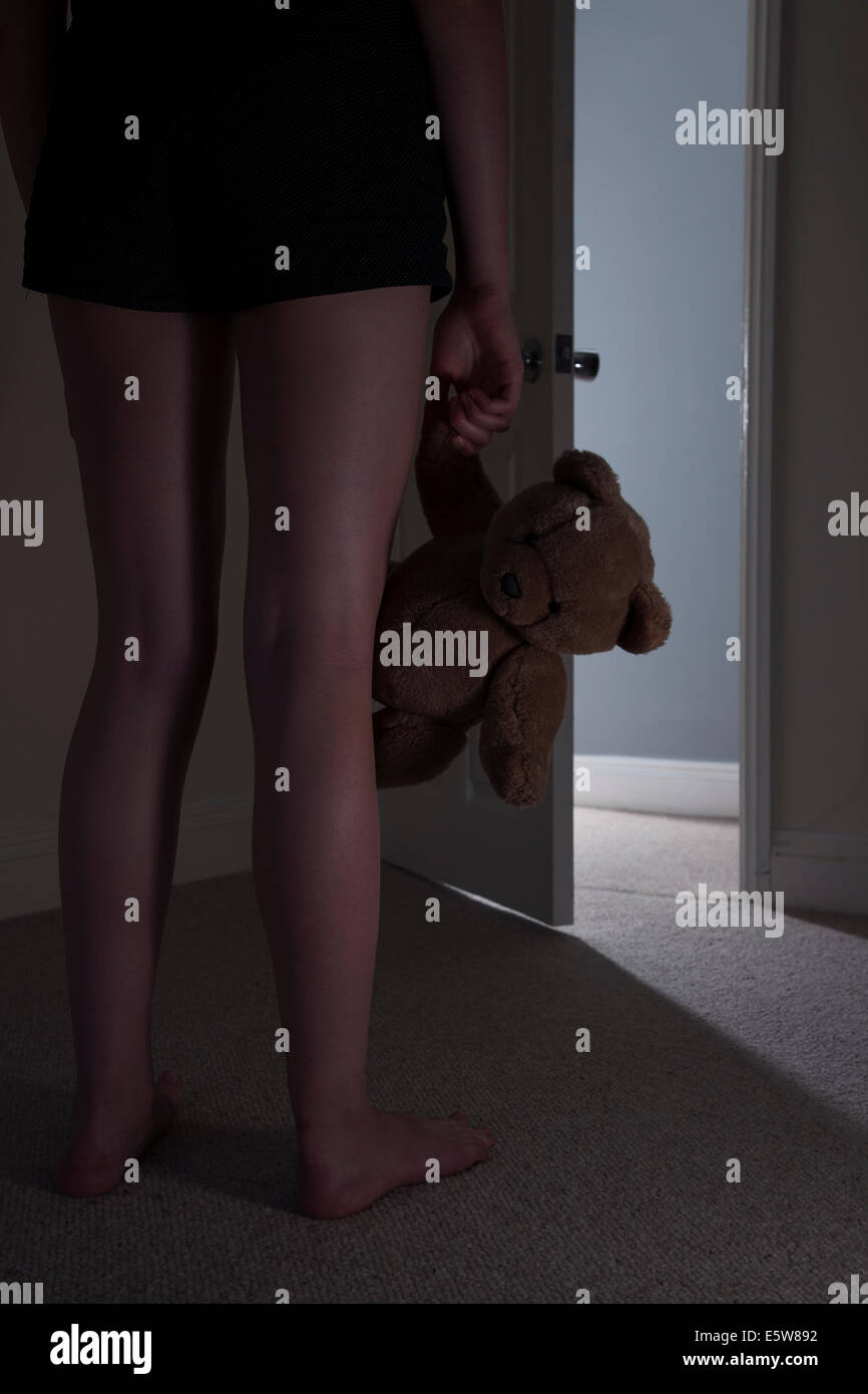 Anonymous young girl standing in front of an open door holding a teddy-bear in a dark room. Stock Photo