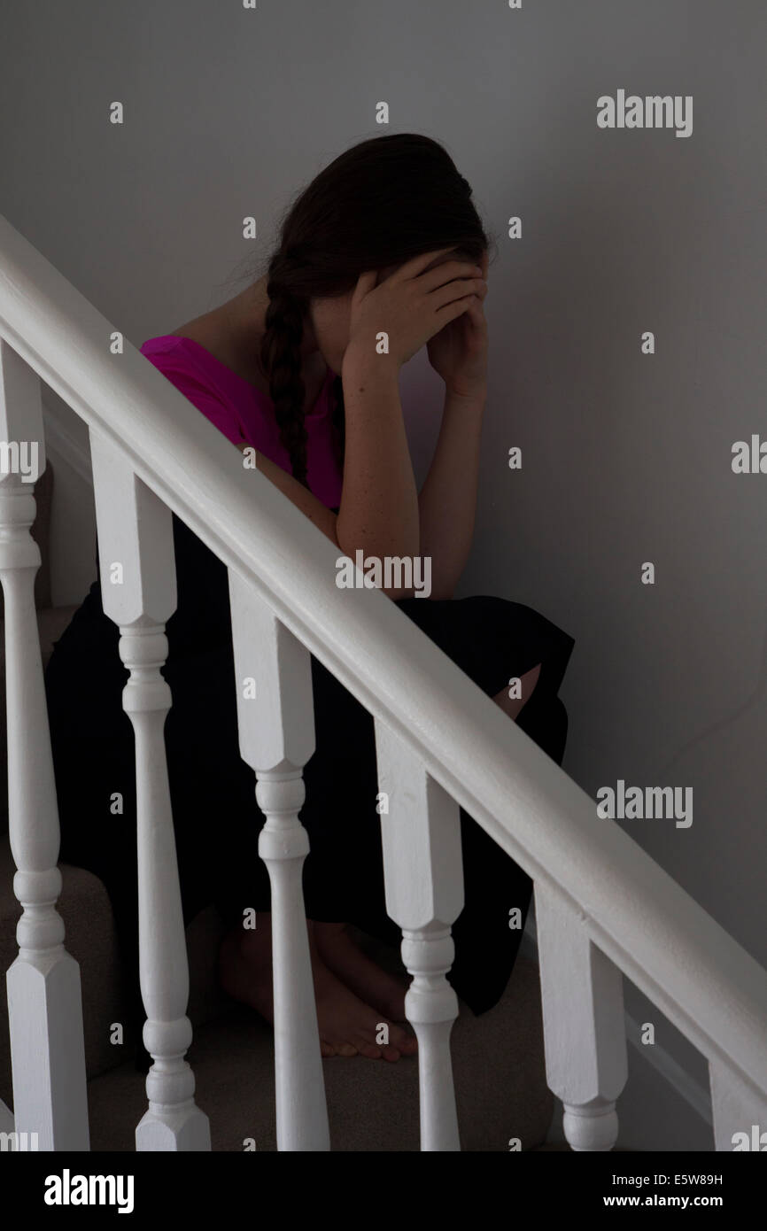 Young female wearing a pink top and hair in plats sitting on the stairs indoors. Stock Photo
