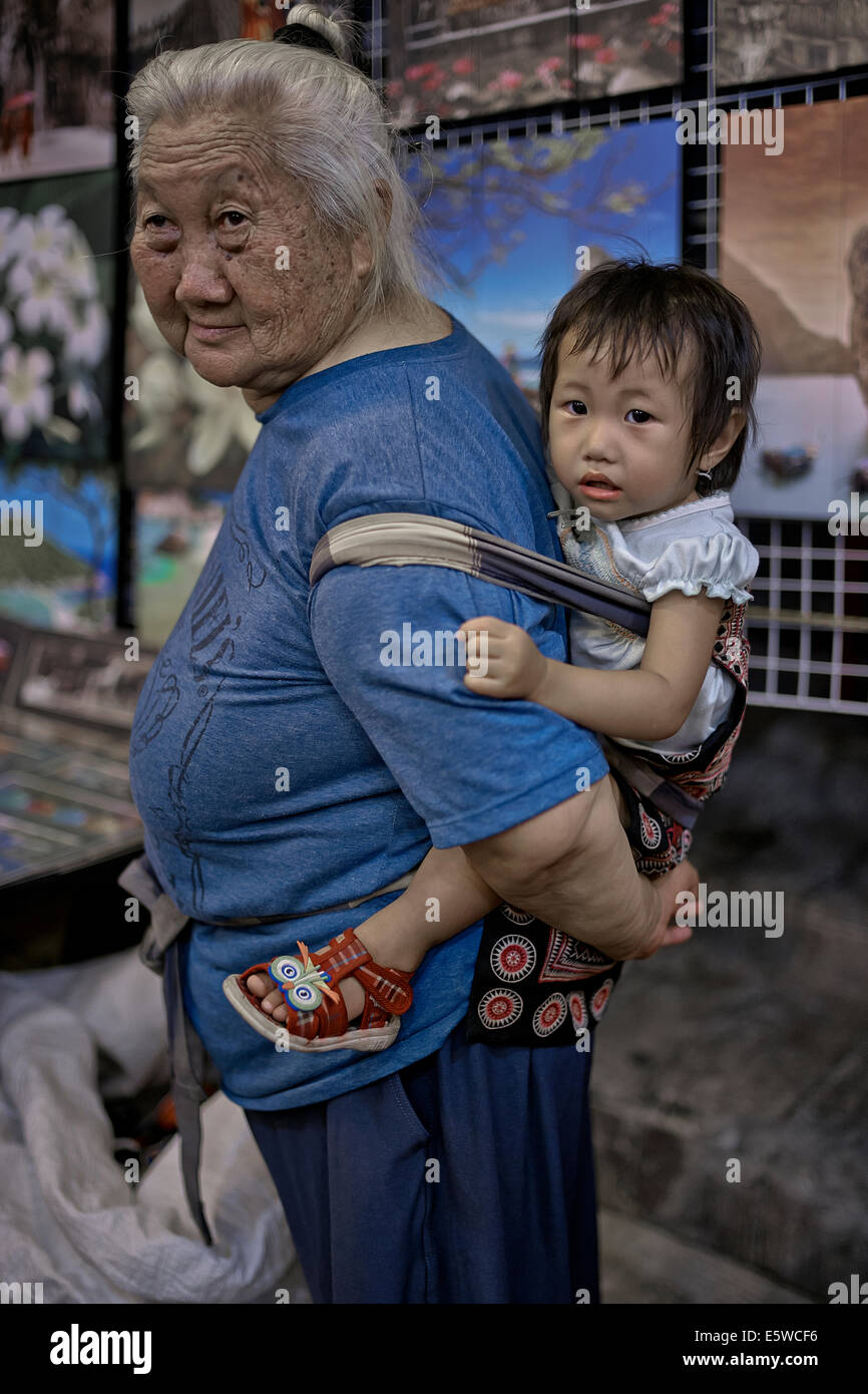 Grandmother carrying infant granddaughter in a back sling harness. Thailand S. E. Asia, Asian grandma carrying child Stock Photo
