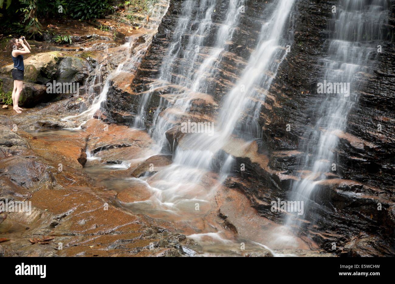 Girl taking photo of waterfall in tropical rainforest region of Borneo. Long exposure photography effect creates silky smooth waterfalls with woman Stock Photo