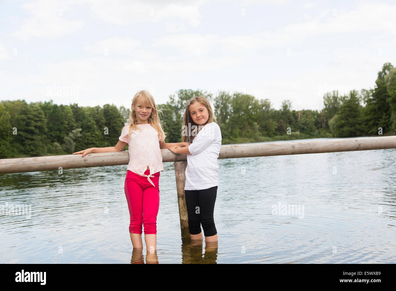 Portrait of two girls holding onto fence whilst paddling in rural lake Stock Photo