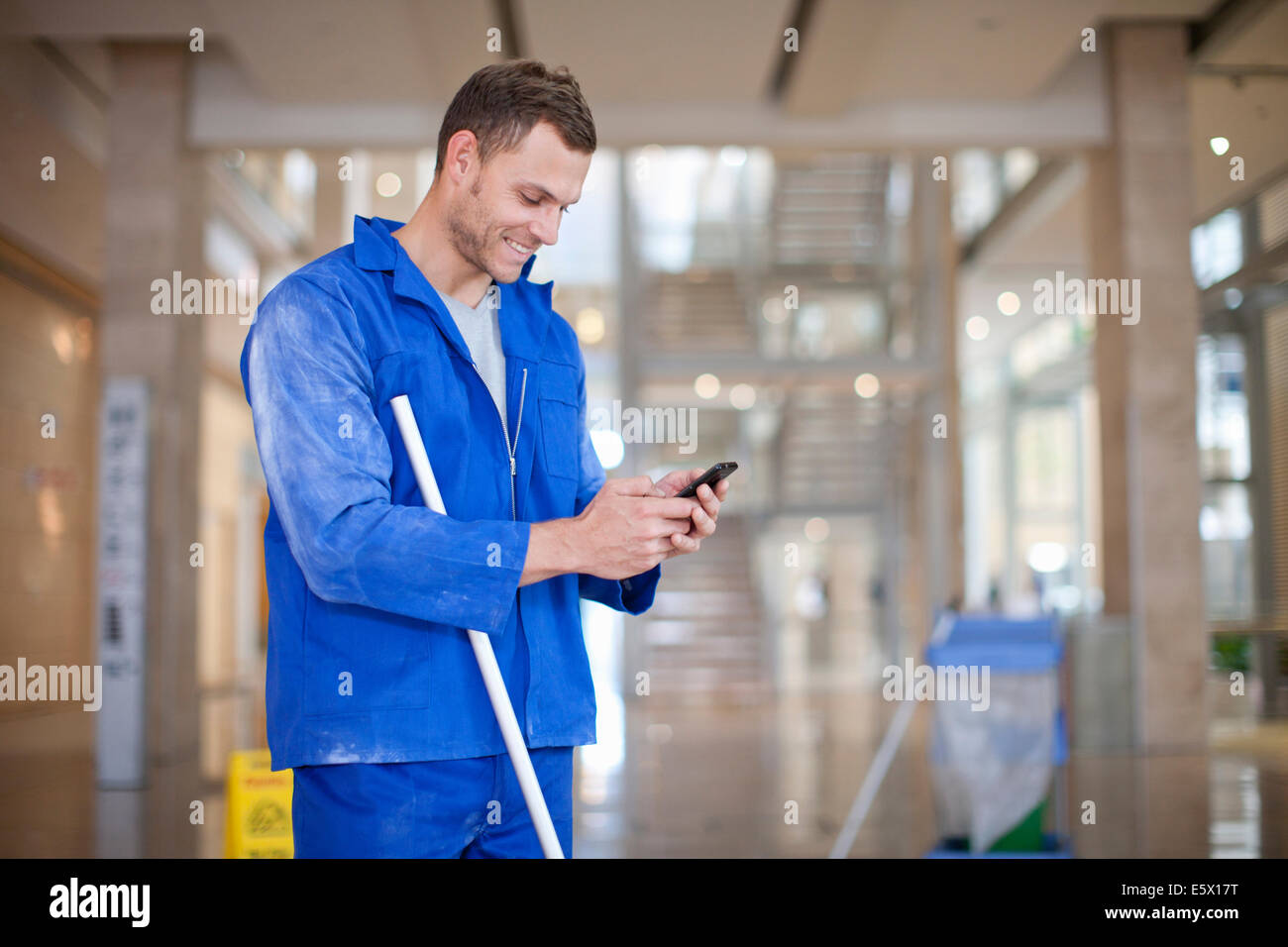 Male cleaner texting on smartphone in office atrium Stock Photo