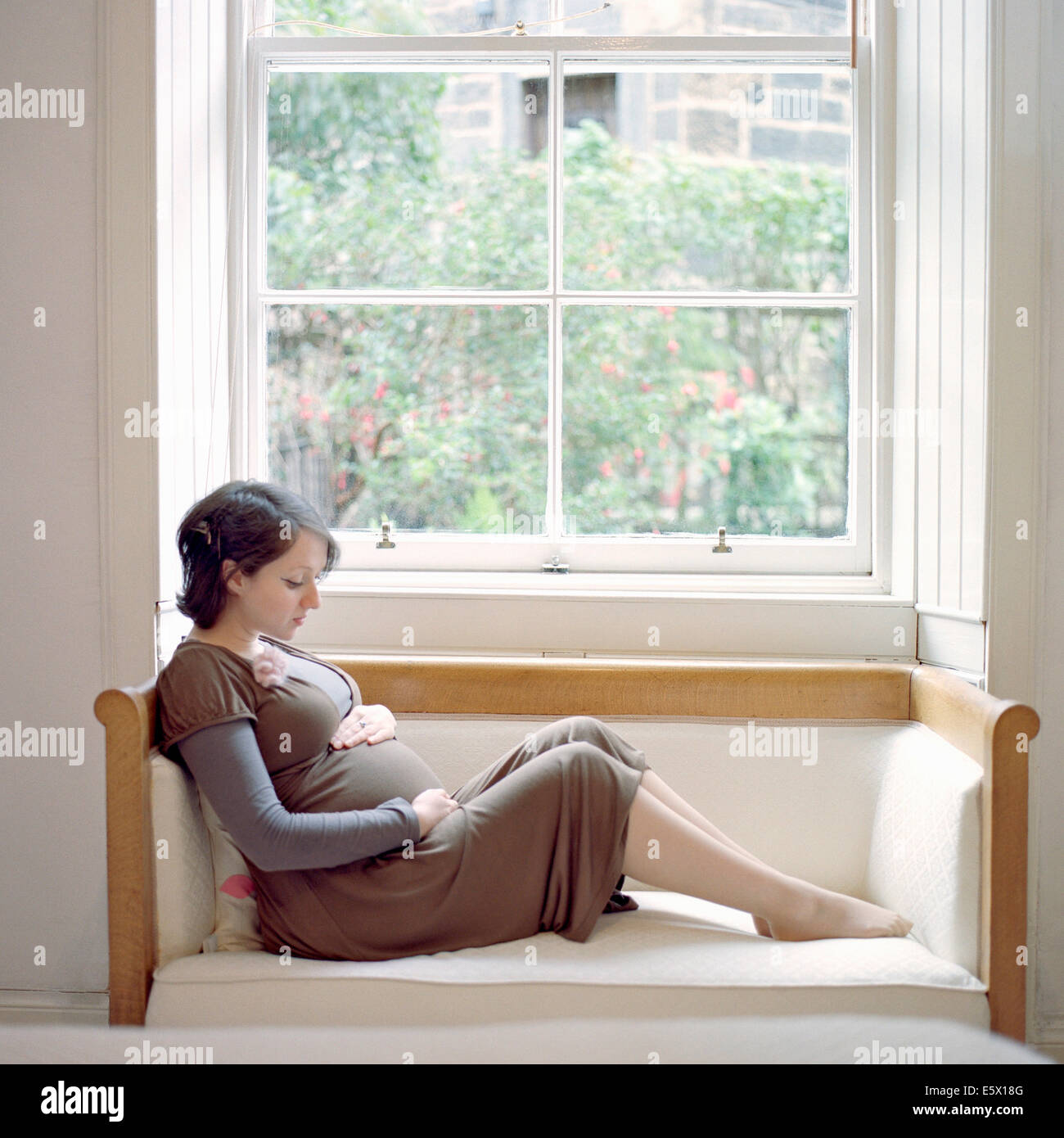 Portrait of mid adult pregnant woman sitting on window seat holding stomach Stock Photo