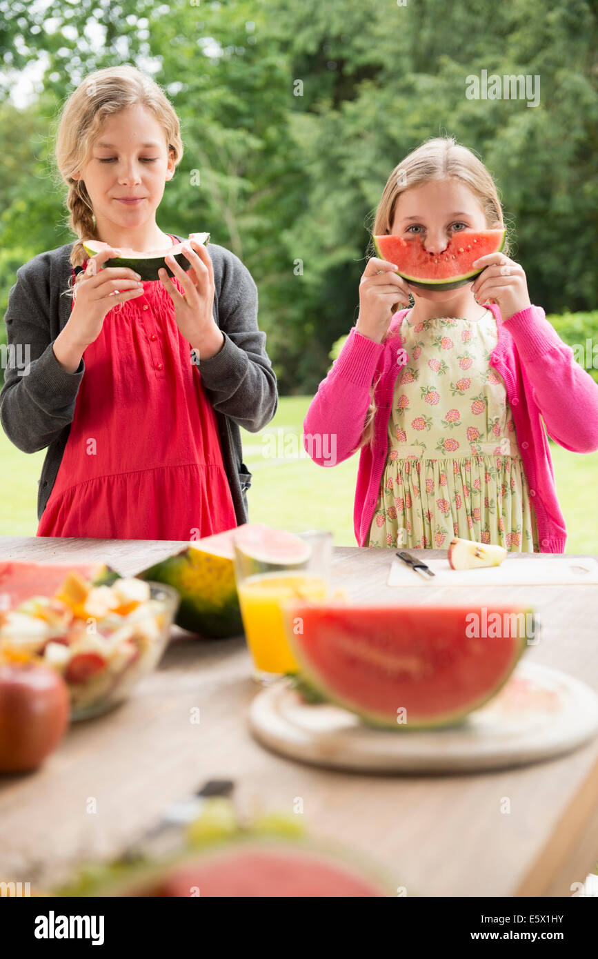 Two sisters at patio table eating and holding up watermelon slice Stock Photo