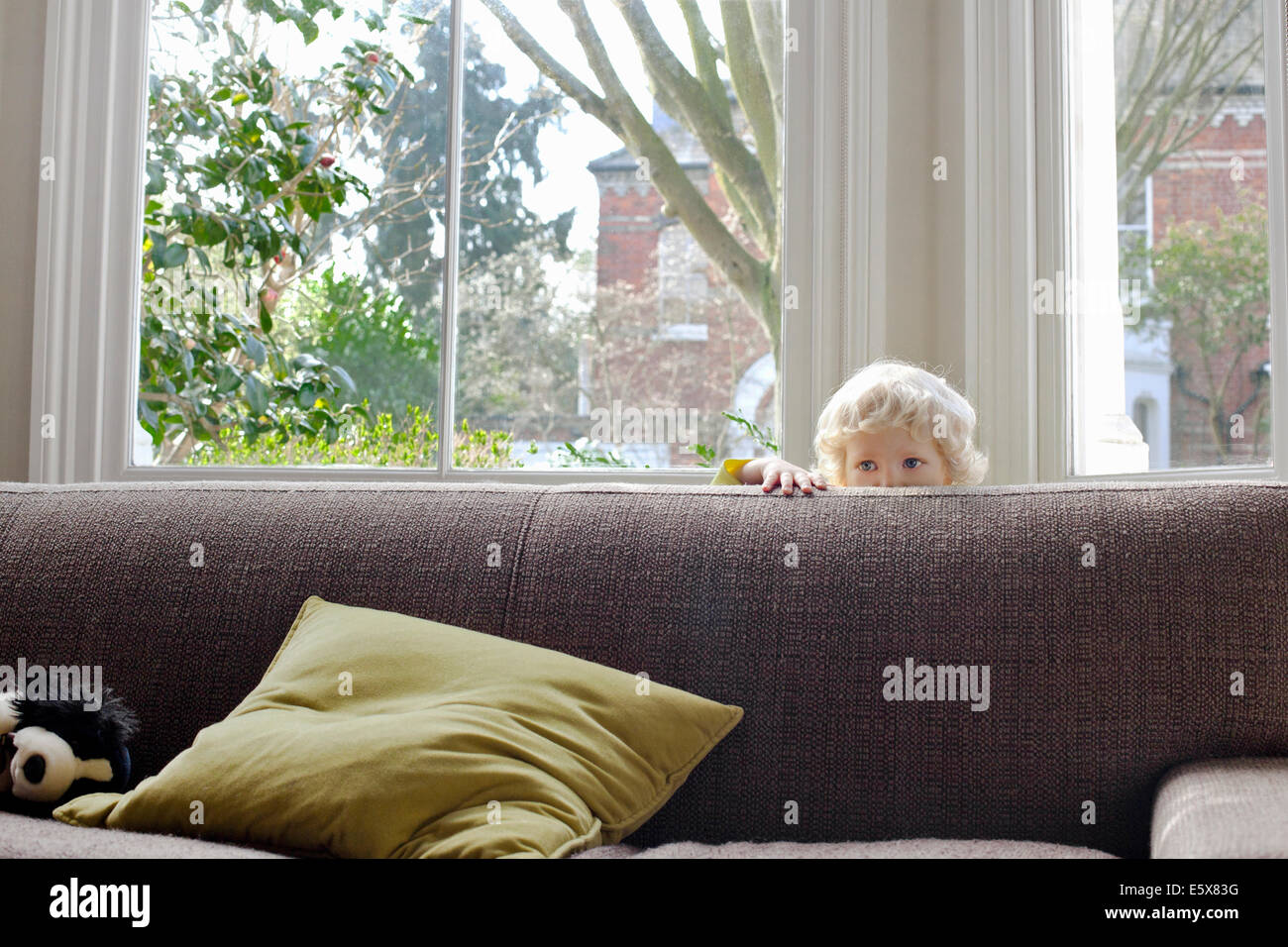 Portrait of cute male blond toddler hiding behind sofa Stock Photo