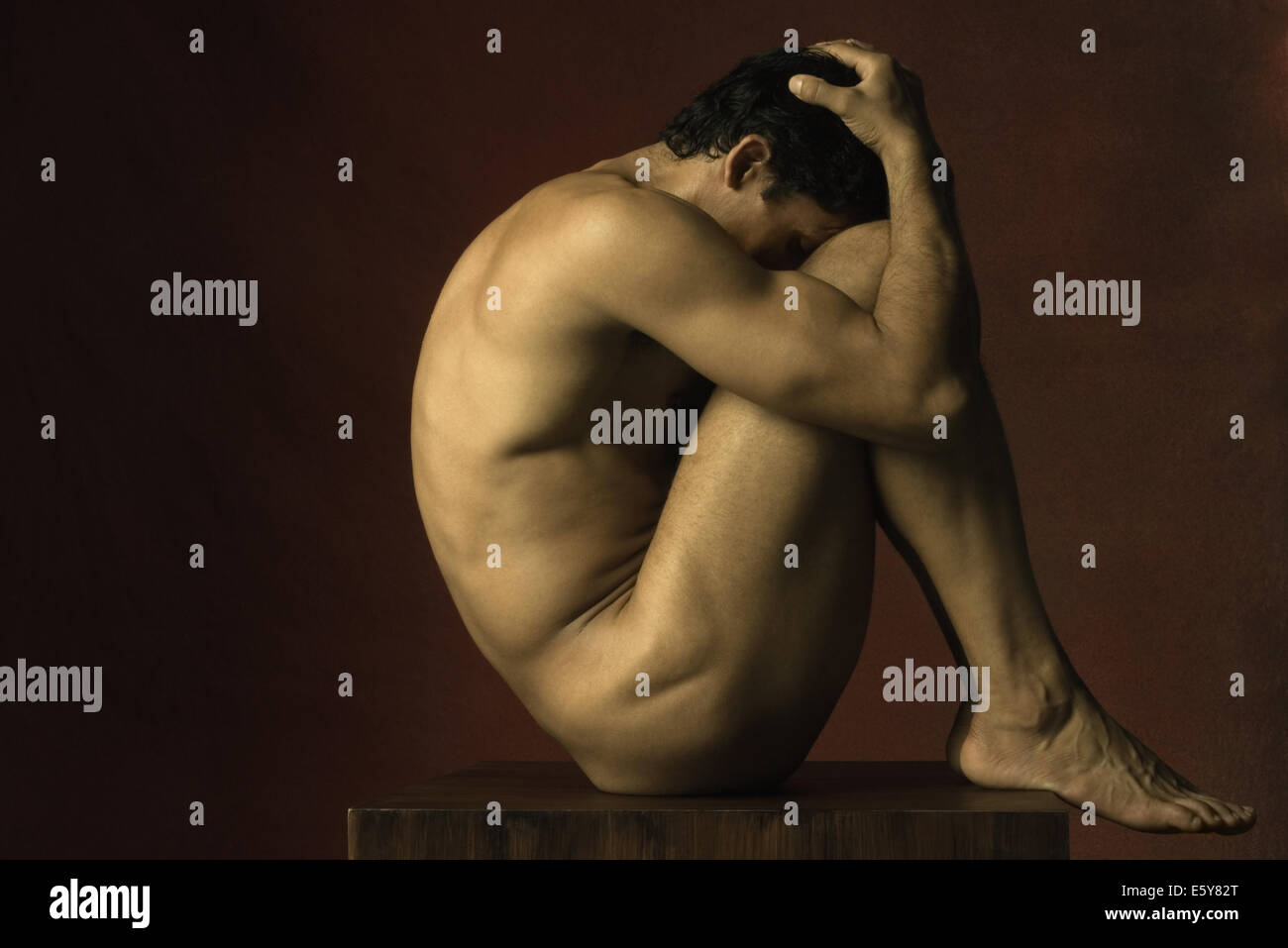 Nude man sitting in fetal position, hands on head, side view Stock Photo