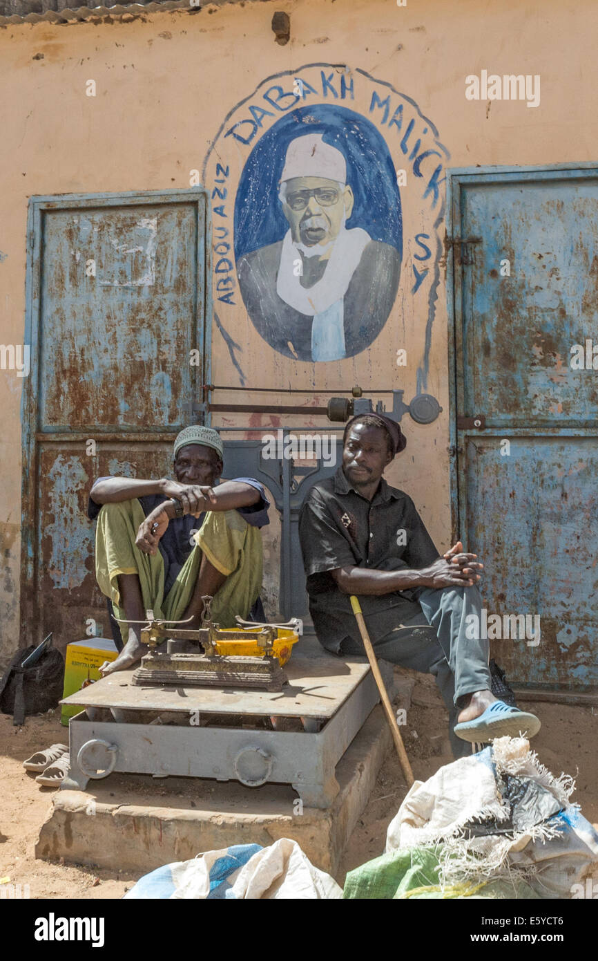Men, sitting on weighing machine, by image of Abdou Aziz Dabakh Malick Sy, 3rd Caliph Tijan Sufi Muslim Brotherhood, Ngoumba village, market, Senegal Stock Photo