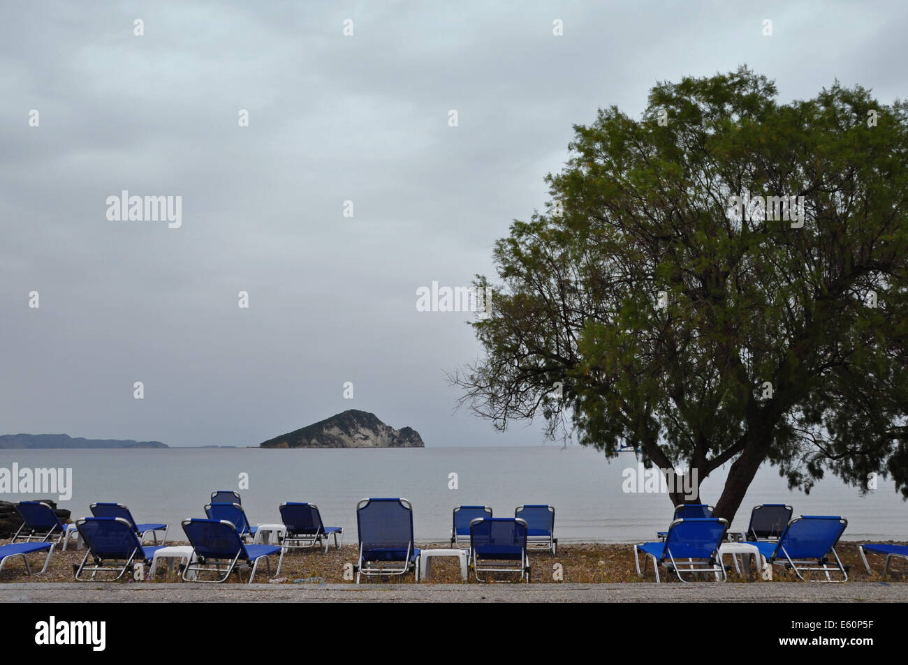 Lounge sunbed chairs with sea view tamarix tree and distant islet, Keri beach, Zakynthos Greece. Stock Photo
