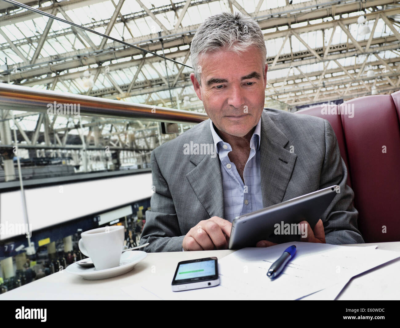 Mature businessman seated at cafe table on railway concourse looking at his iPad tablet computer with iPhone and notes in foreground Stock Photo