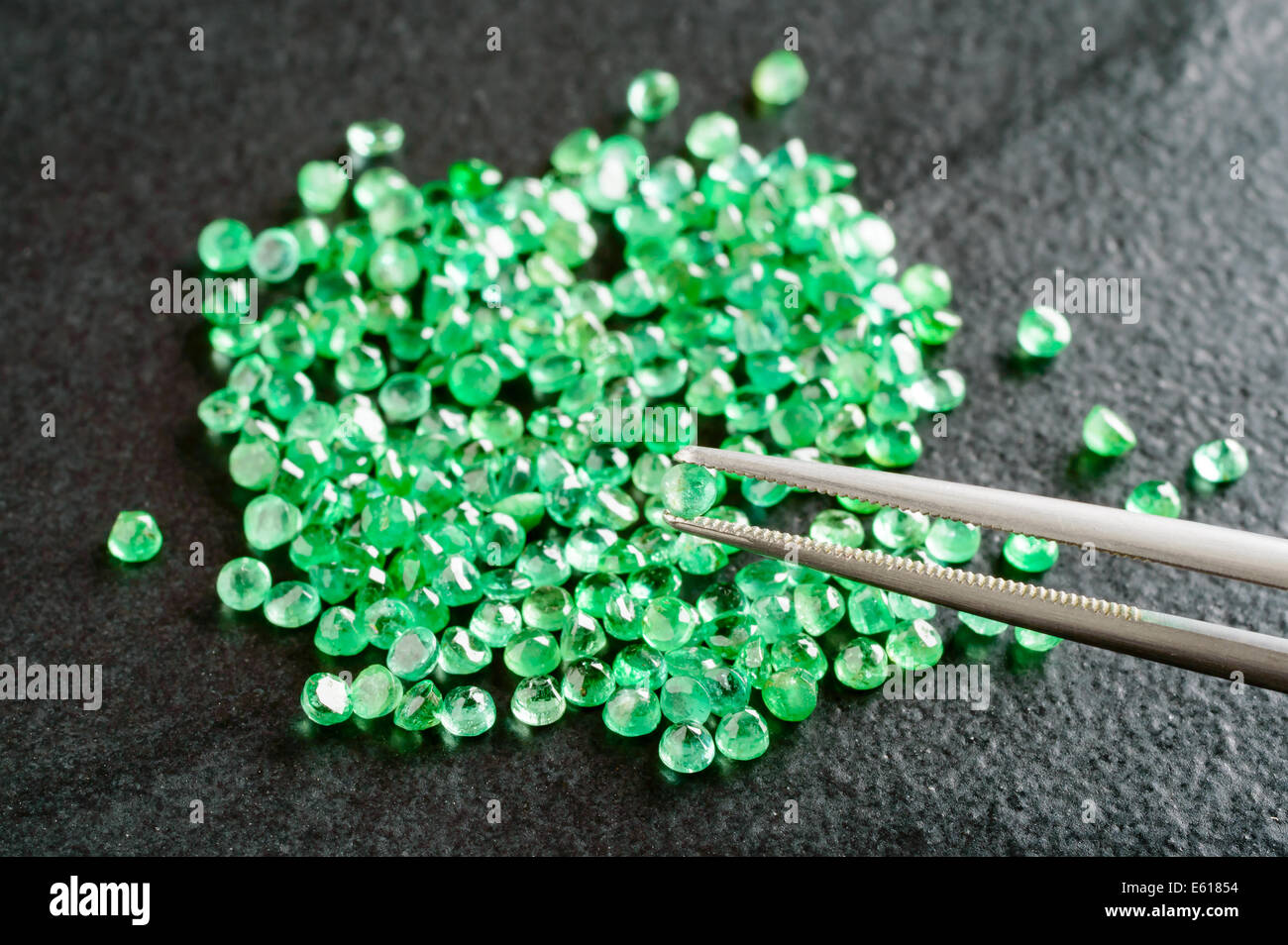 Pile of green, round cut emeralds on black stone plate. One emerald held by tweezers. Stock Photo
