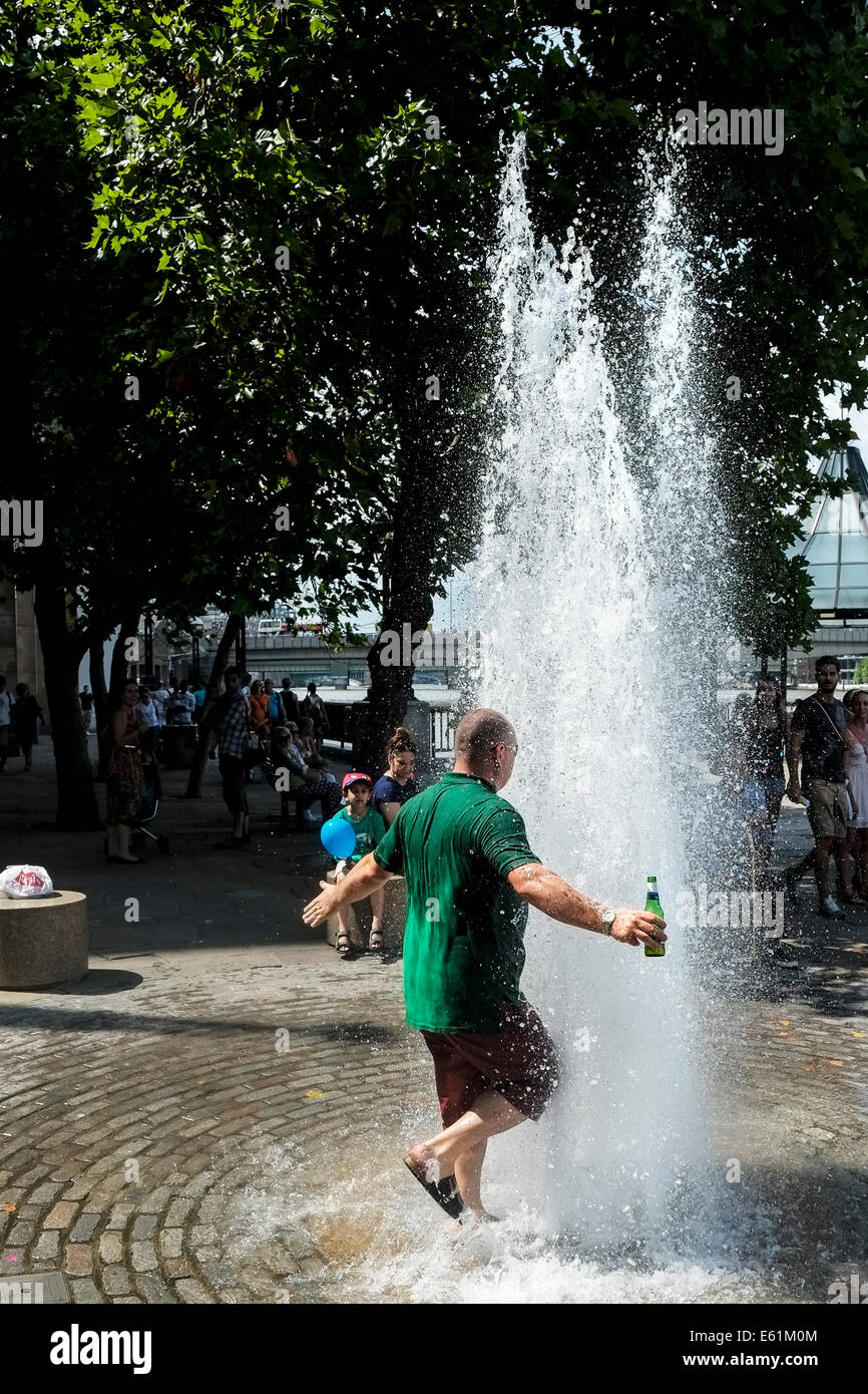 A man holding a bottle of beer walking into a fountain of water to escape the hot weather in London. Stock Photo
