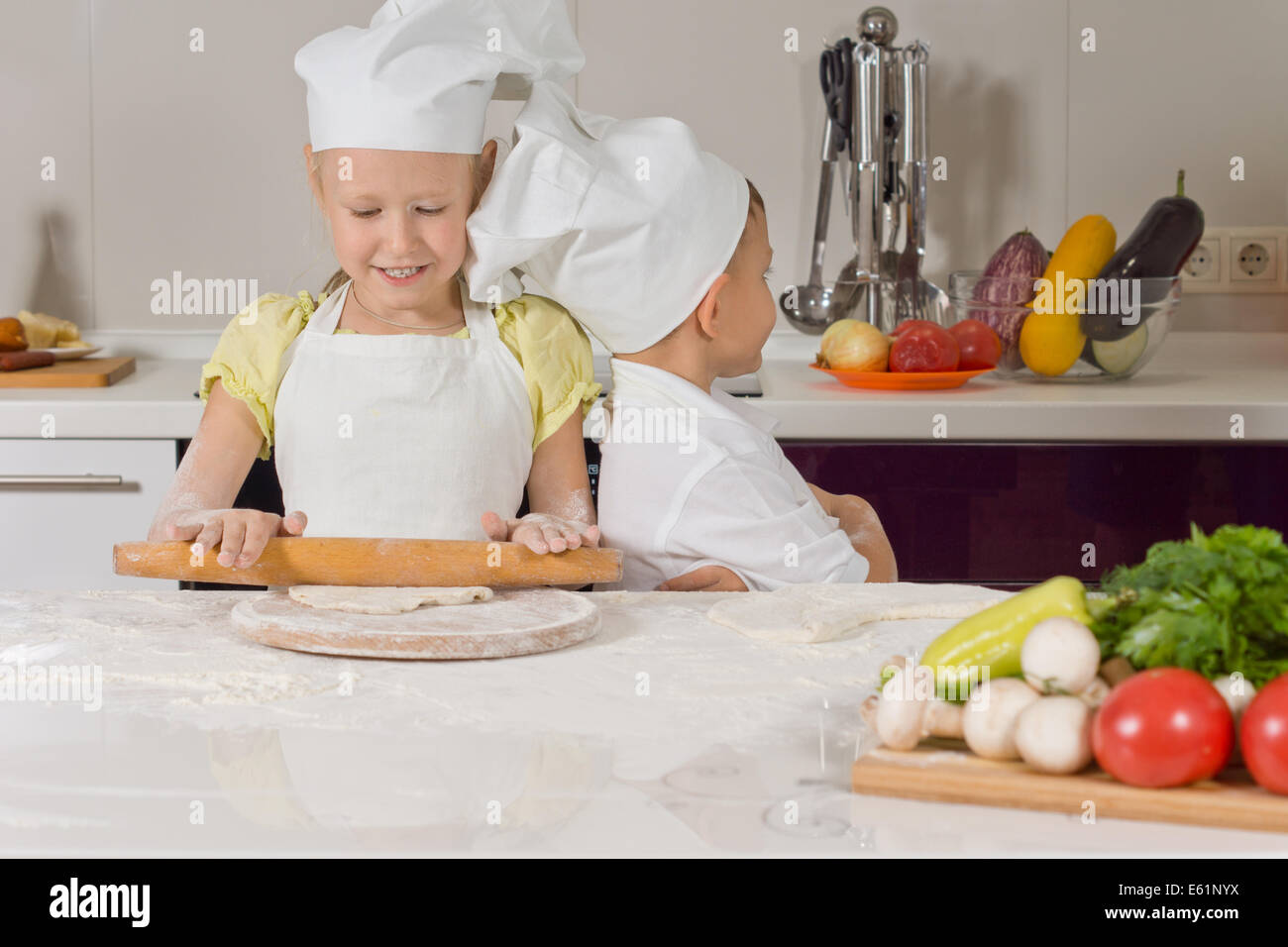 Cute little girl learning to bake in her cooks uniform standing at the kitchen counter rolling out dough with a rolling pin Stock Photo