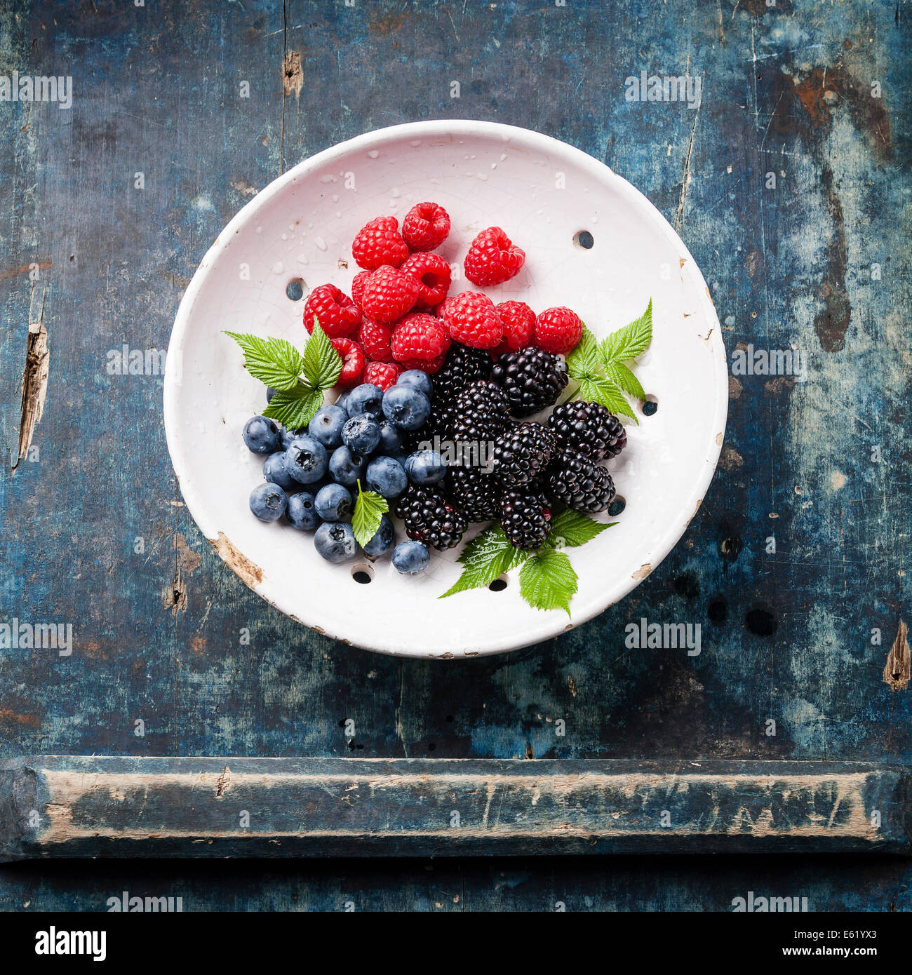 Mix of fresh berries with leaves in vintage ceramic colander on blue wooden background Stock Photo