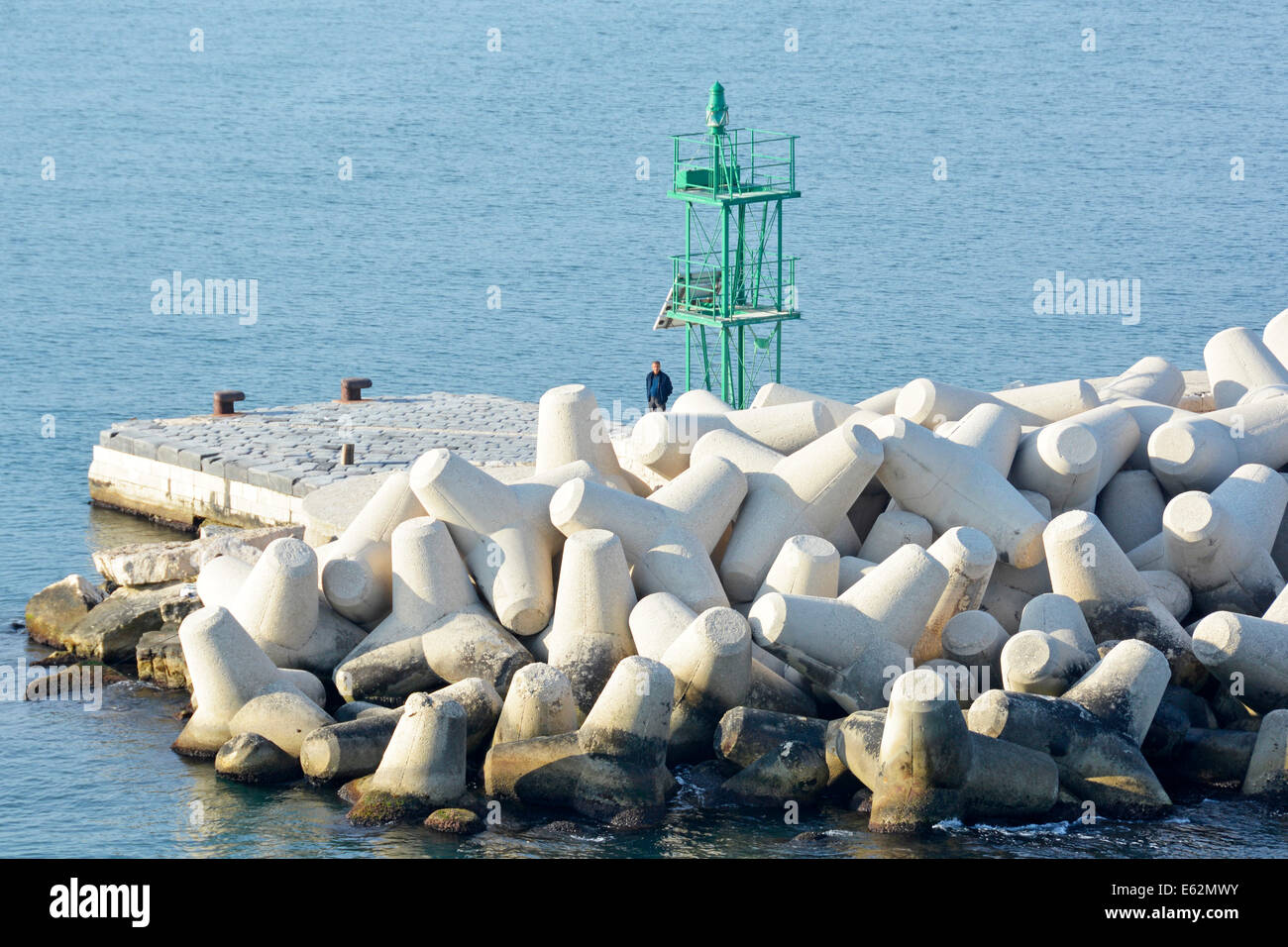 Man standing beside Dolosse type precast concrete blocks in breakwater protect green channel marker on harbour wall Bari Port Puglia Italy Adriatic Stock Photo