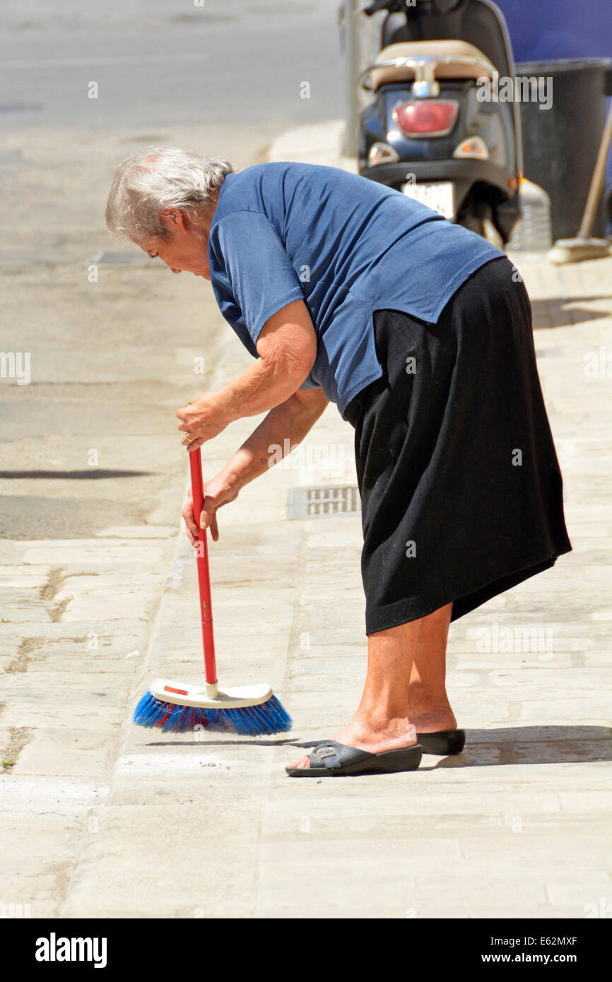 Street scene old woman senior citizen bending to sweep pavement outside her home in Polignano a Mare town in Province of Bari Apulia Puglia Italy  EU Stock Photo