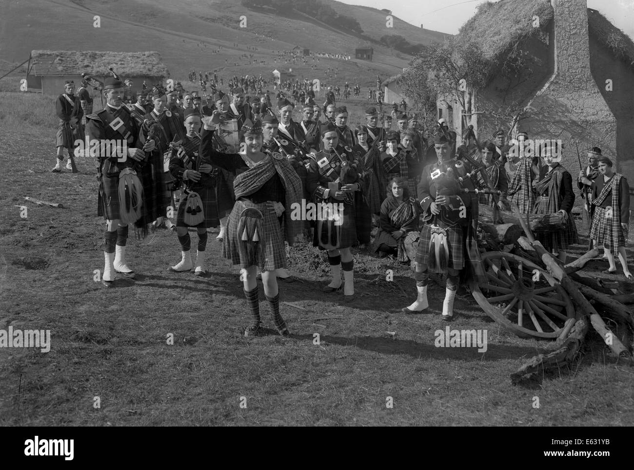 1920s SILENT MOVIE STILL WOMAN IN KILT LEADING BAND OF SCOTSMEN IN UNIFORMS PLAYING BAGPIPES PAST STONE COTTAGES IN HIGHLANDS Stock Photo