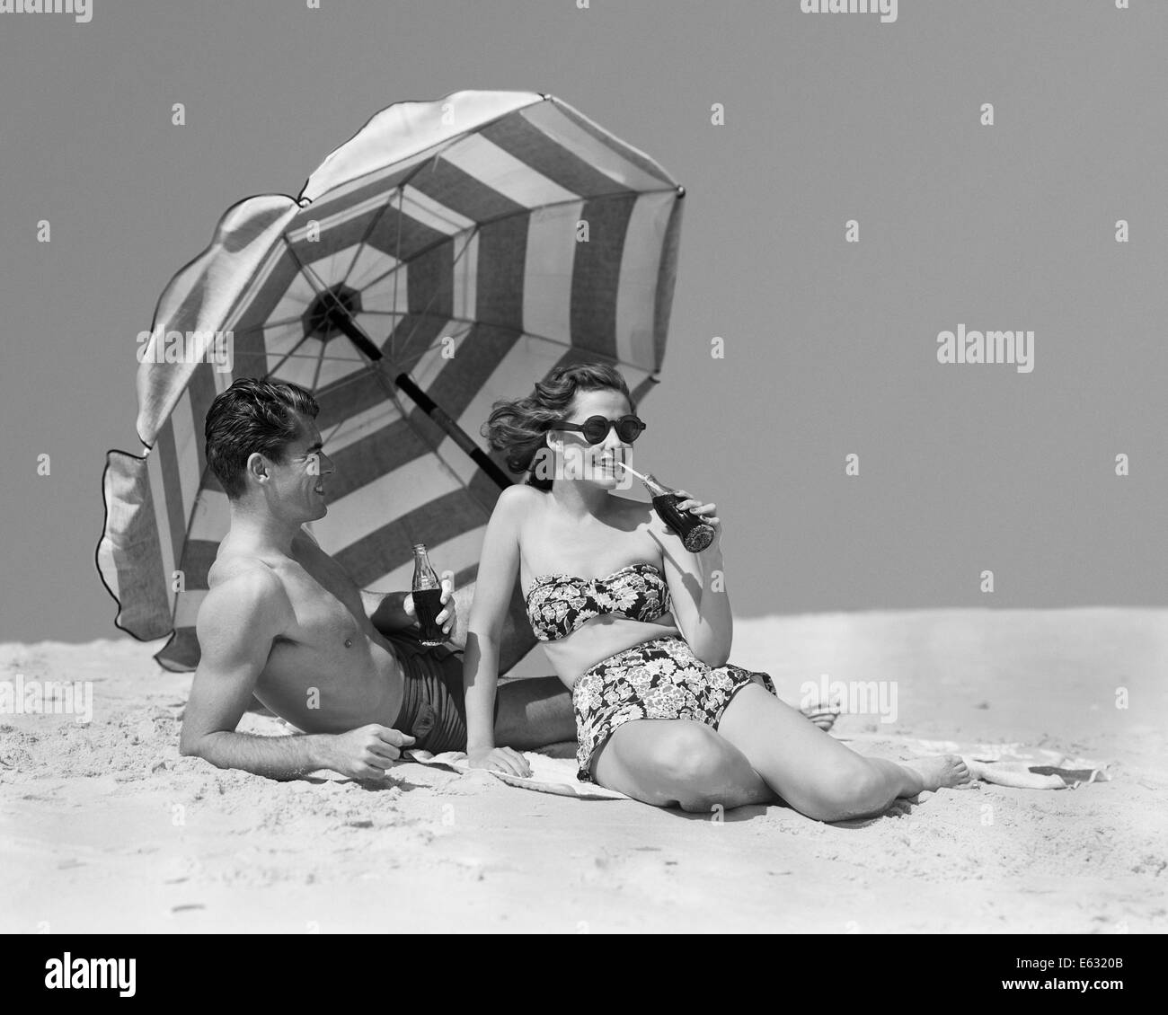 1950s COUPLE MAN WOMAN SUNBATHING DRINKING BOTTLES SODA BY BEACH UMBRELLA Stock Photo
