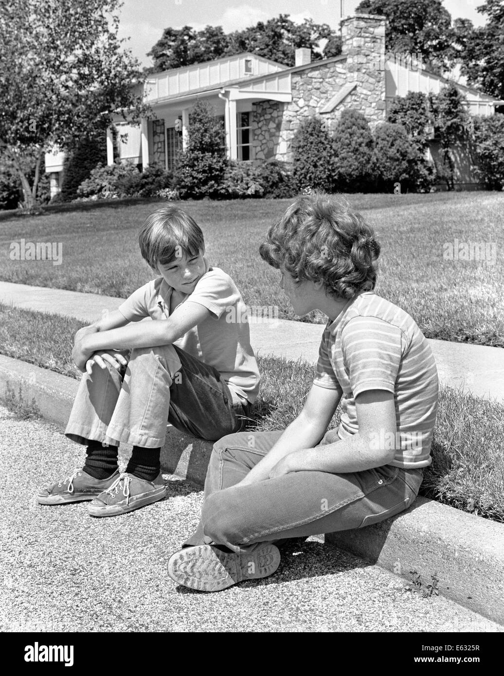 1970s TWO EARLY TEENAGE BOYS SITTING ON CURB SUBURBAN NEIGHBORHOOD TALKING Stock Photo