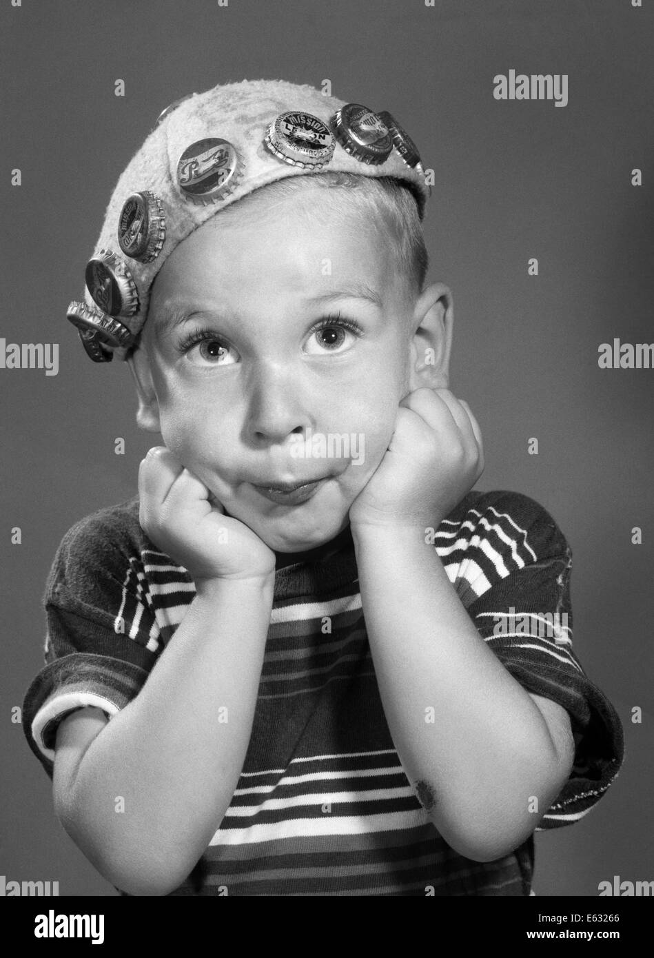 1950s PORTRAIT OF BOY RESTING HEAD ON FISTS LOOKING AT CAMERA WEARING BEANIE HAT DECORATED WITH SODA BOTTLE CAPS Stock Photo