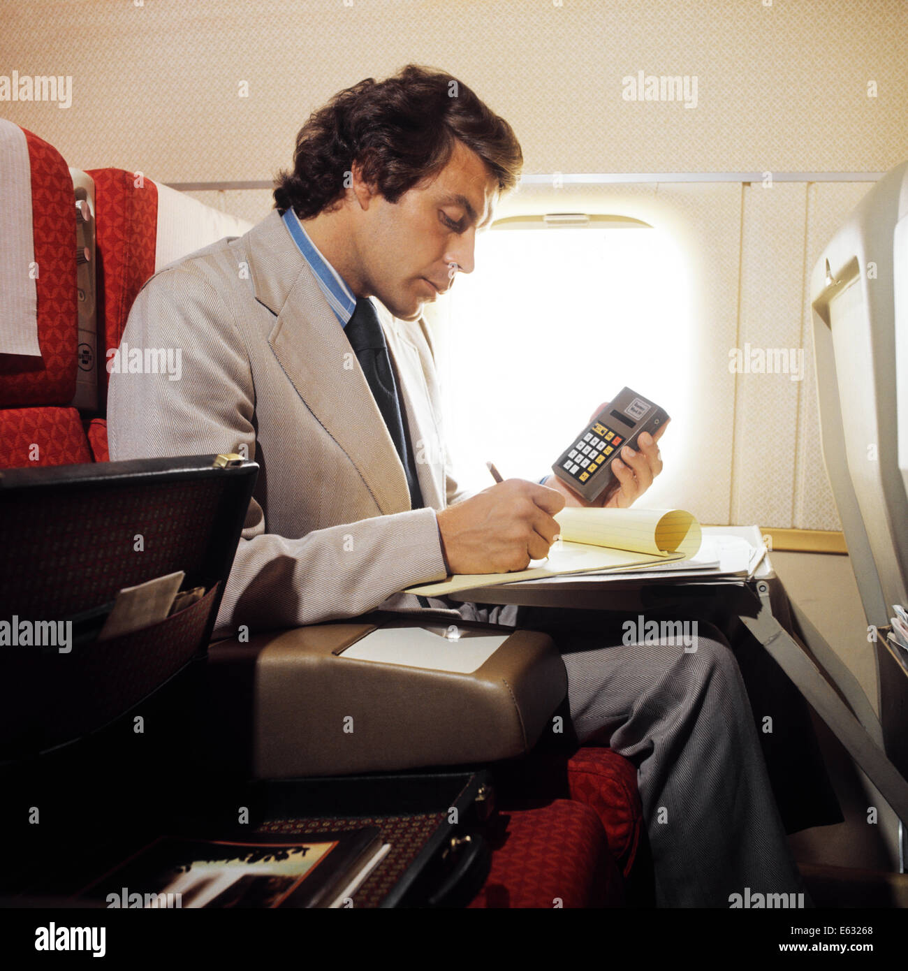 1970s 1980s BUSINESS MAN ABOARD PLANE WORKING ON PAPERS HOLDING CALCULATOR IN HAND Stock Photo