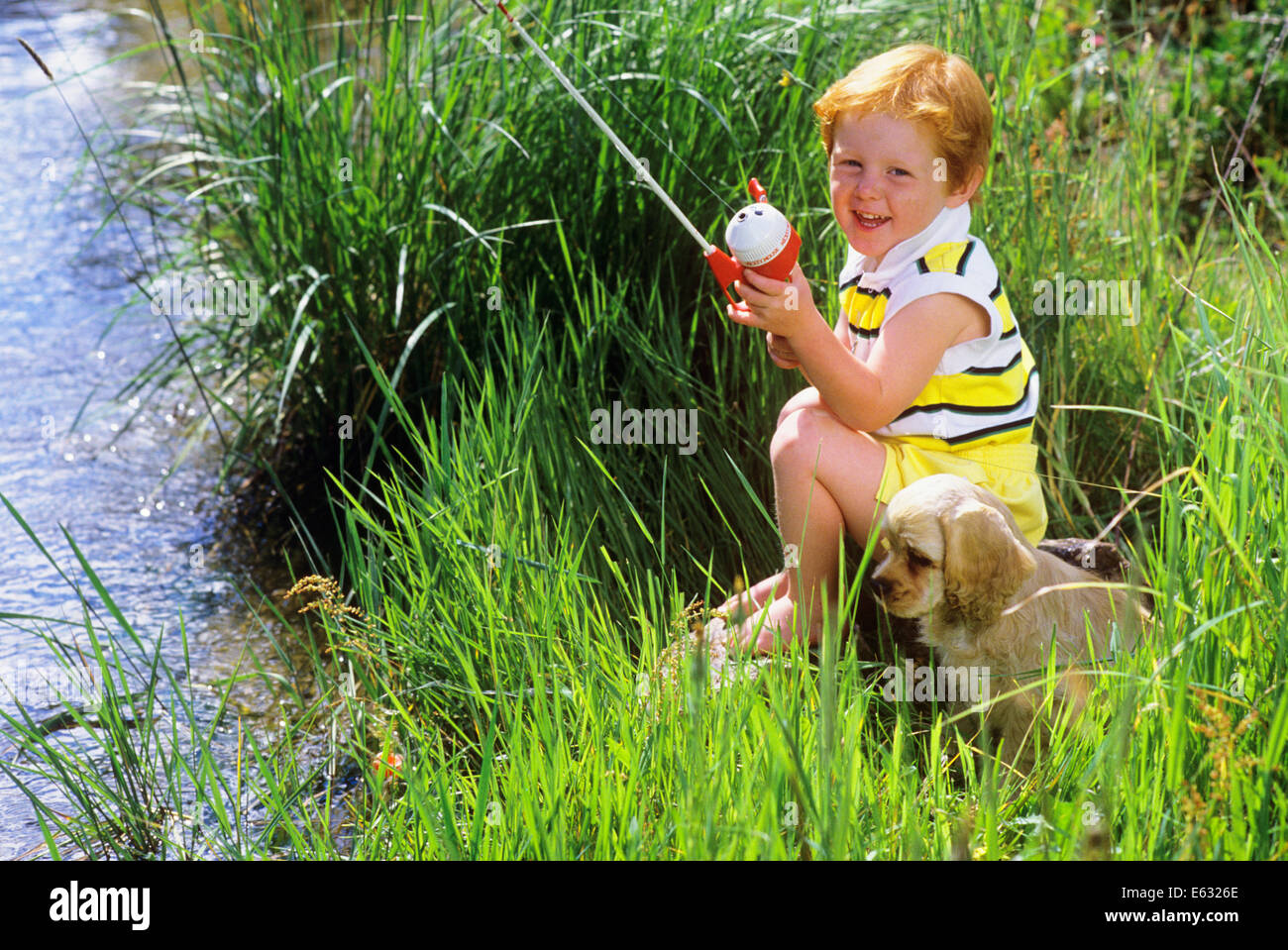 REDHEADED BOY FISHING FROM BANK WITH COCKER SPANIEL BESIDE HIM Stock Photo