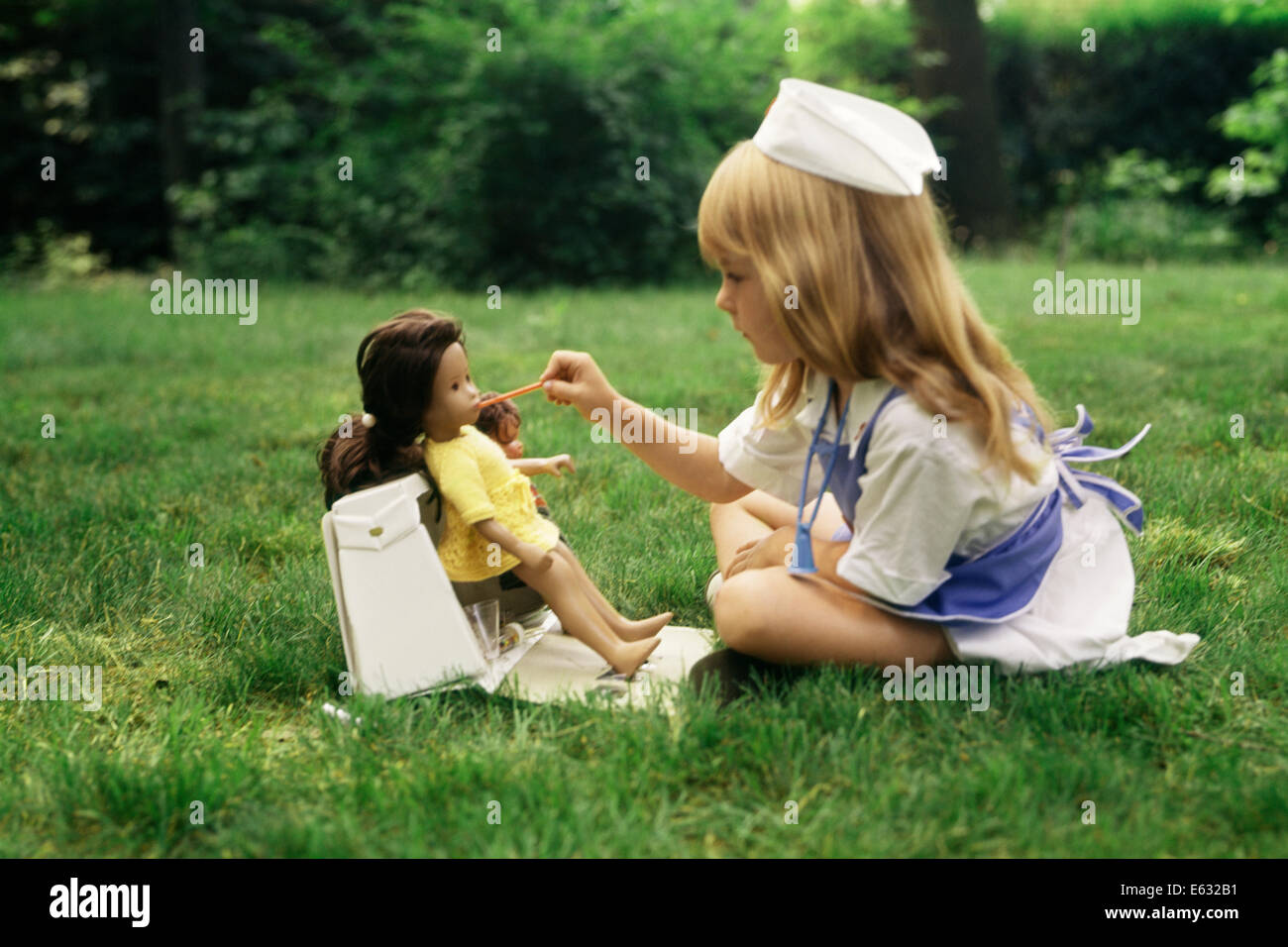 1970s LITTLE GIRL IN NURSE OUTFIT TAKING TEMPERATURE OF DOLL PLAYING ROLE PRACTICE LAWN YARD Stock Photo