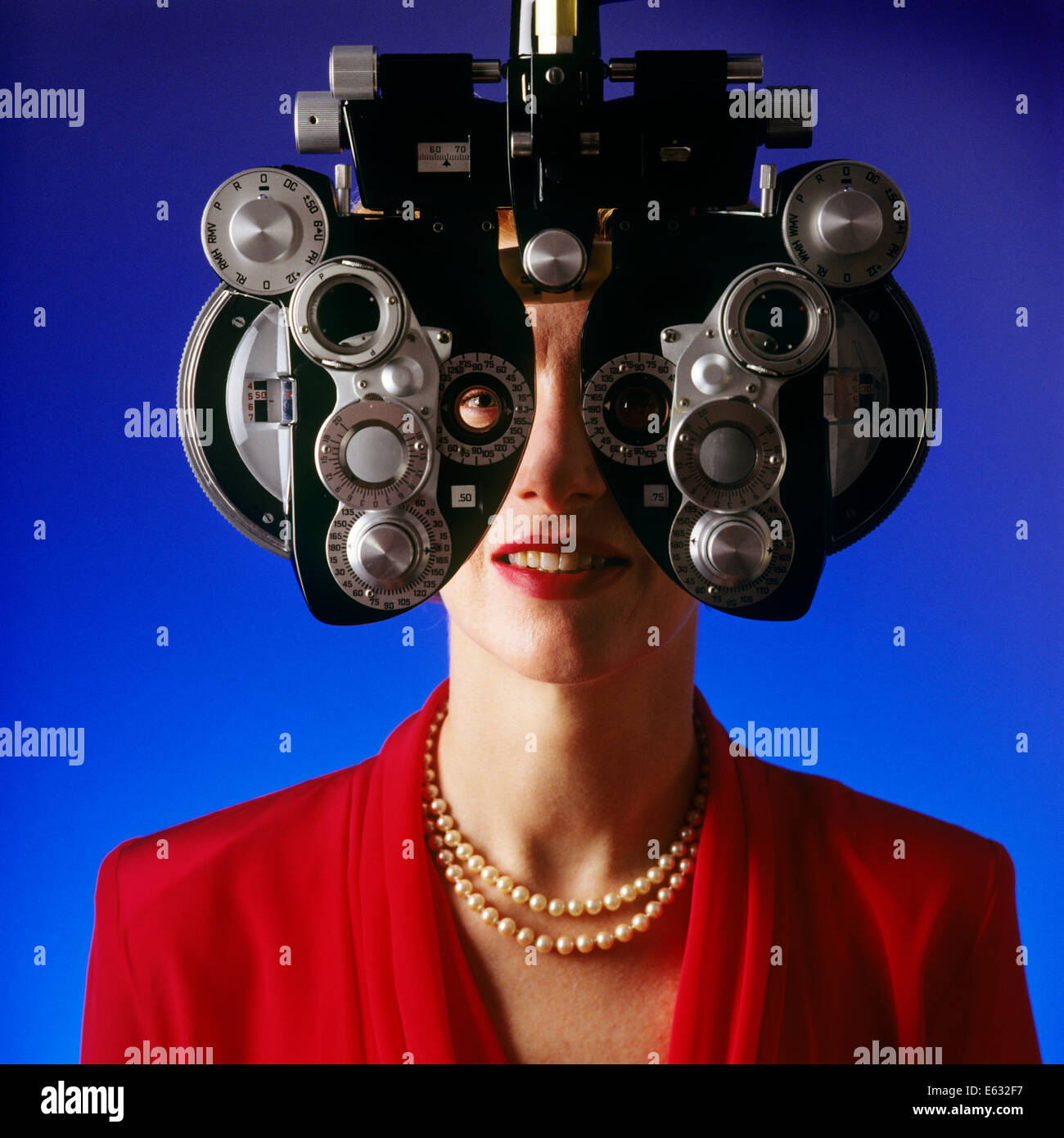 WOMAN RED BLOUSE PEARLS RECEIVING EYE EXAM LOOKING THROUGH PHOROPTER Stock Photo