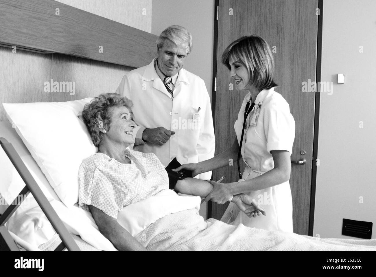 1980s ELDERLY WOMAN IN HOSPITAL BED HAVING BLOOD PRESSURE TAKEN BY FEMALE NURSE MALE DOCTOR STANDING BESIDE HER Stock Photo