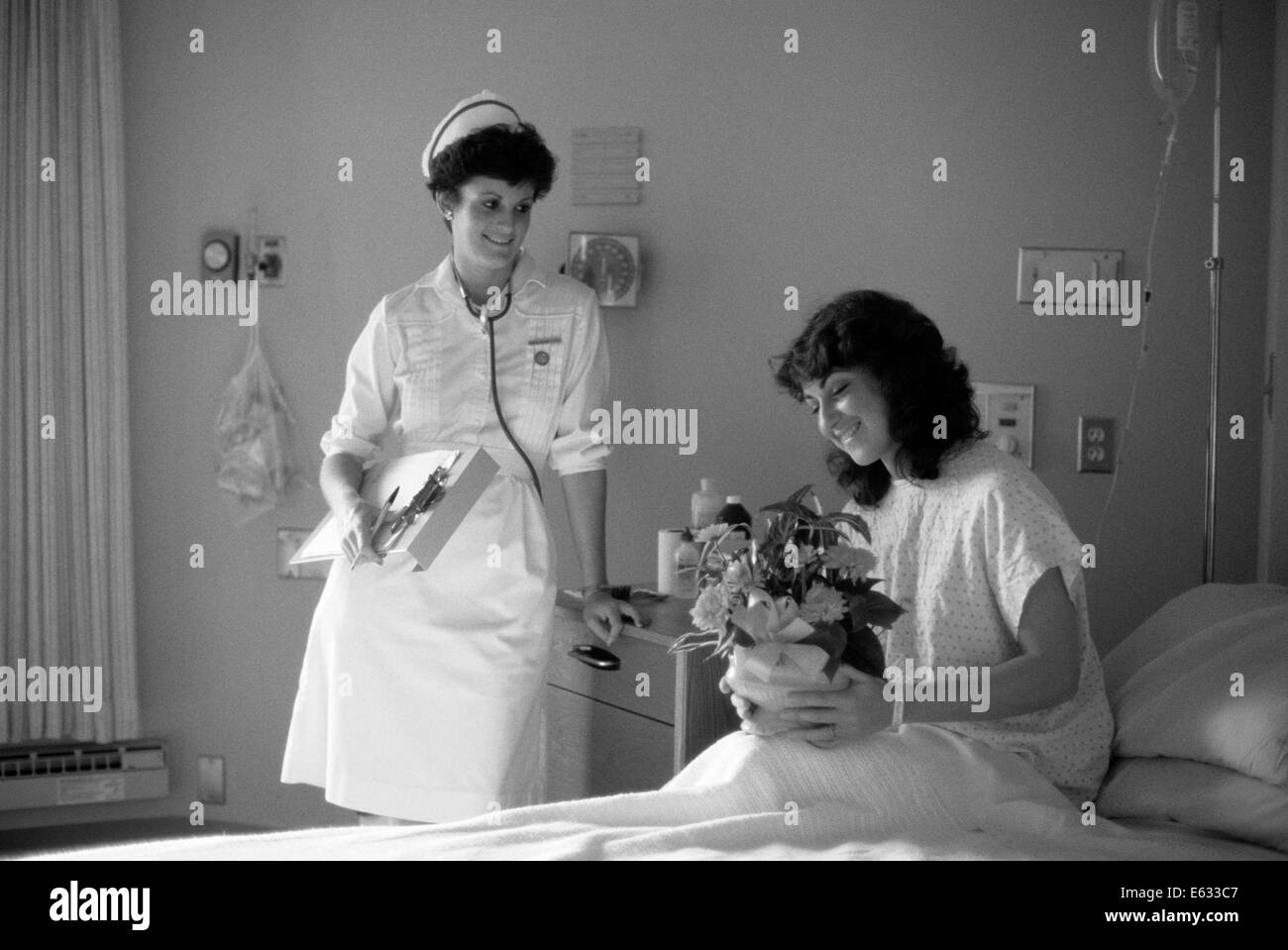 1980s SMILING FEMALE PATIENT SITTING UP IN HOSPITAL BED HOLDING FLOWER ARRANGEMENT SMILING FEMALE NURSE HOLDING CHART LOOKING ON Stock Photo