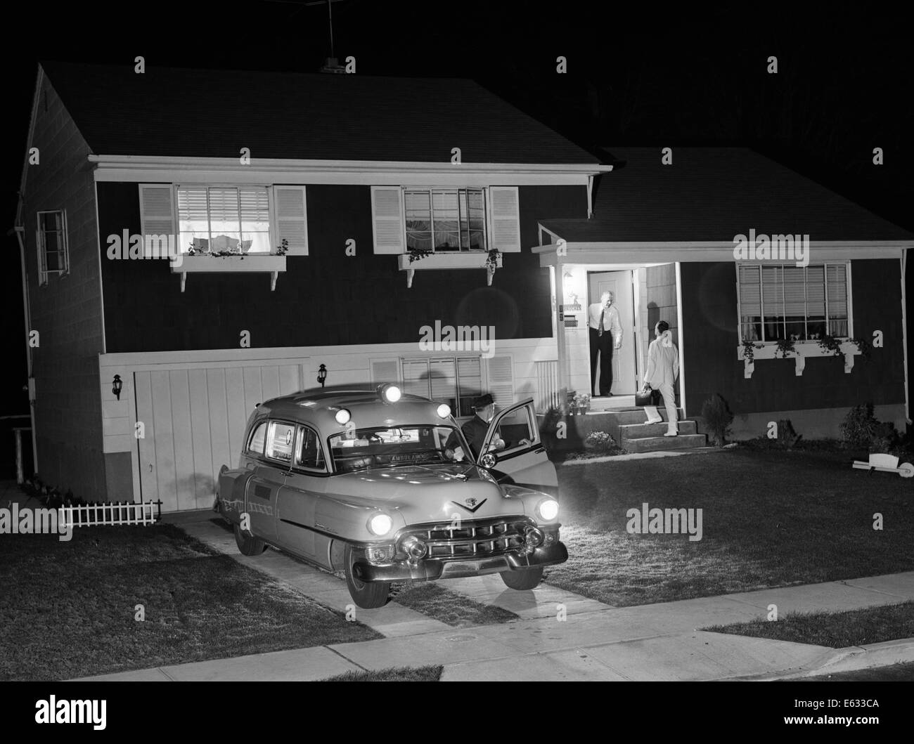 1950s AMBULANCE AT NIGHT PARKED IN SUBURBAN HOME DRIVEWAY WITH DOOR OPEN WHILE EMT DRIVER TALKS TO RESIDENT AT HOUSE DOOR Stock Photo