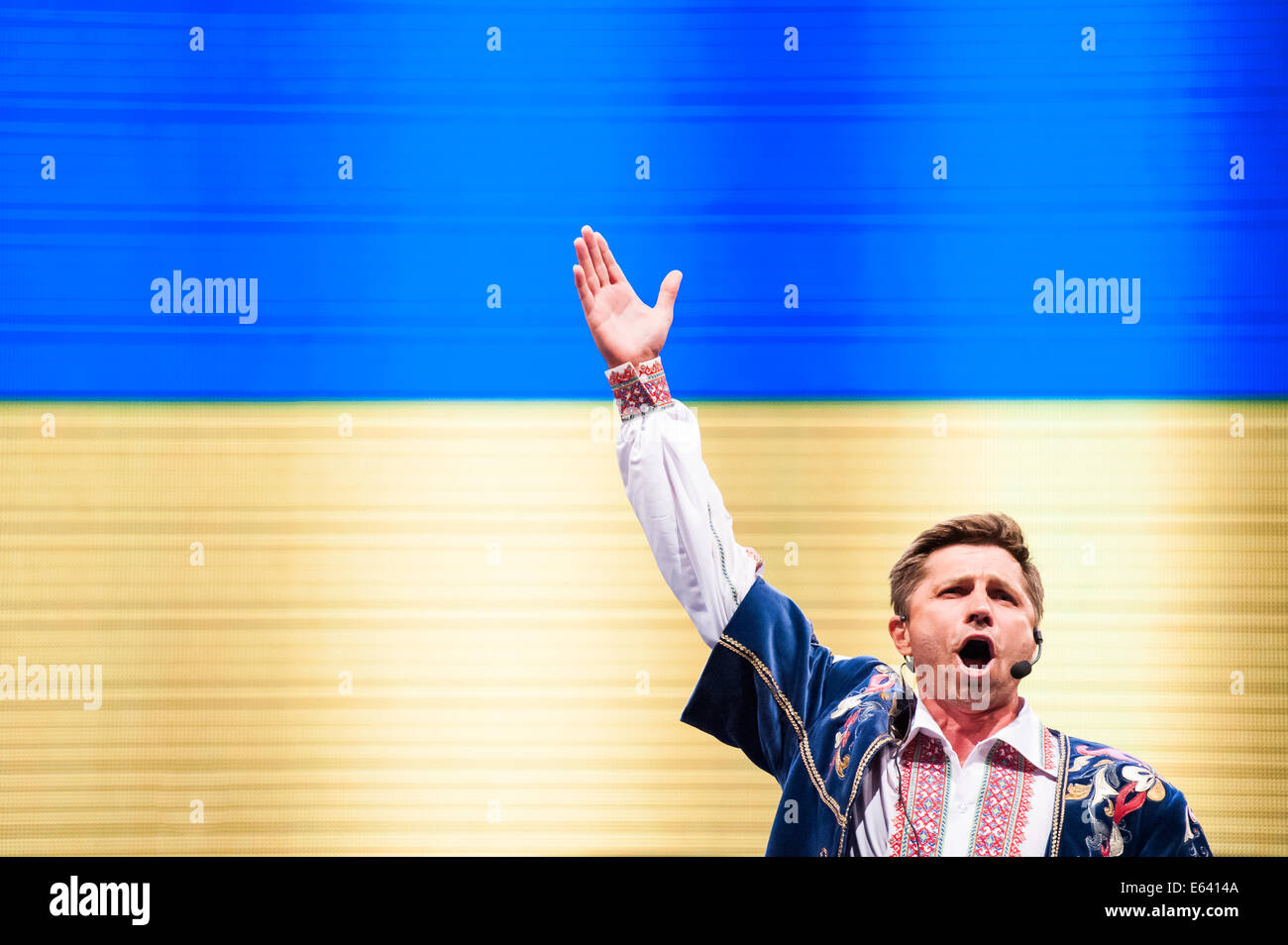 A Ukrainian folk singer with his right hand in the air, with Ukrainian flag in the background, performing at Folkart festival Stock Photo