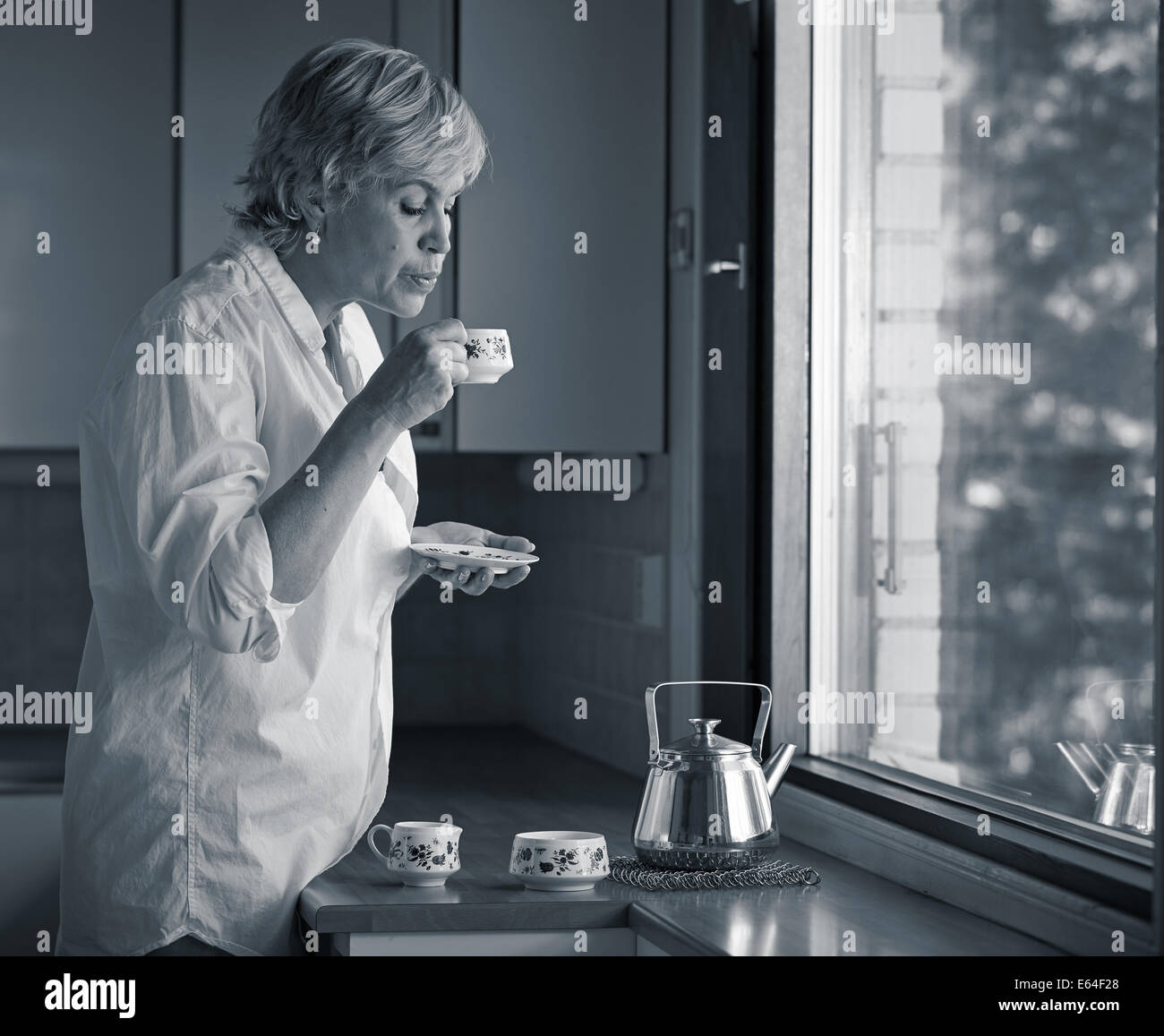 Serene morning moment, mature woman drinking coffee in the kitchen, sunlight coming from the window Stock Photo