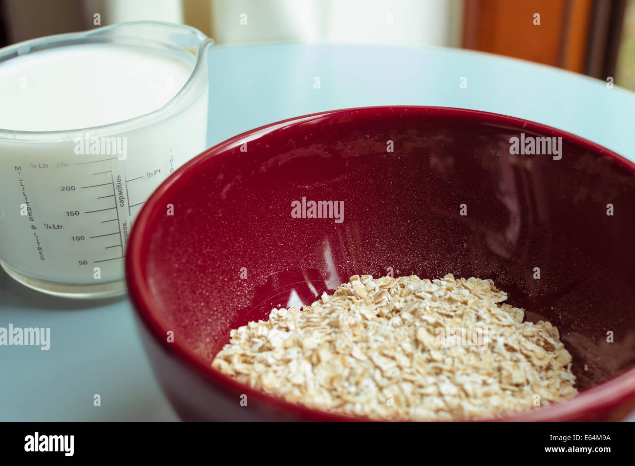porridge oats in a red bowl; milk-filled measuring jug in the background on a blue table-top. Stock Photo
