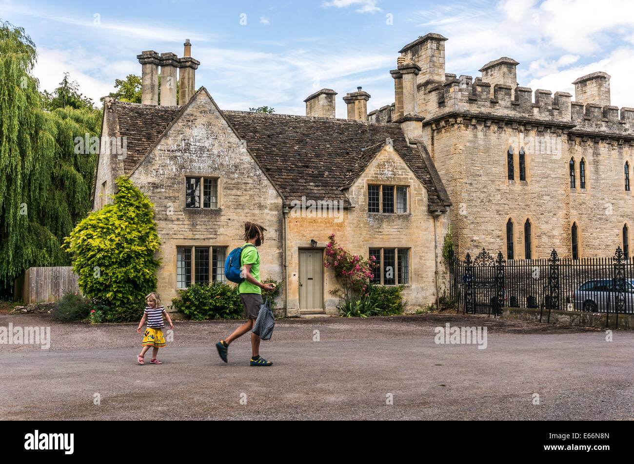 Man / father and child, walking past old house next to Cecily Hill barracks (faux medieval castle), Cirencester Park entrance, Cotswolds, England, UK. Stock Photo