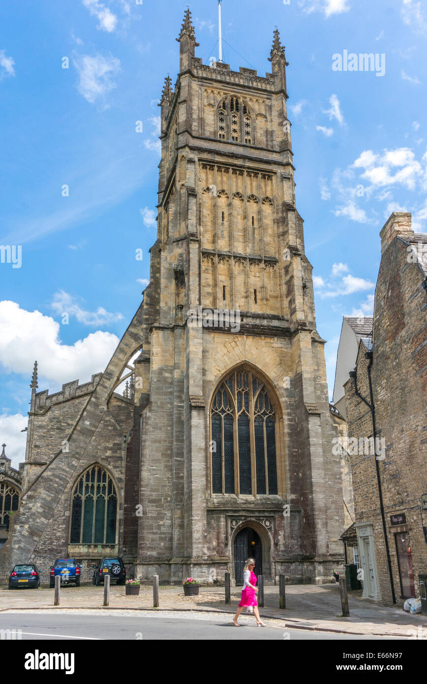 Woman in a pink dress walking past the landmark Church of St John Baptist, Cirencester town centre, Cotswolds, Gloucestershire, England, UK. Stock Photo