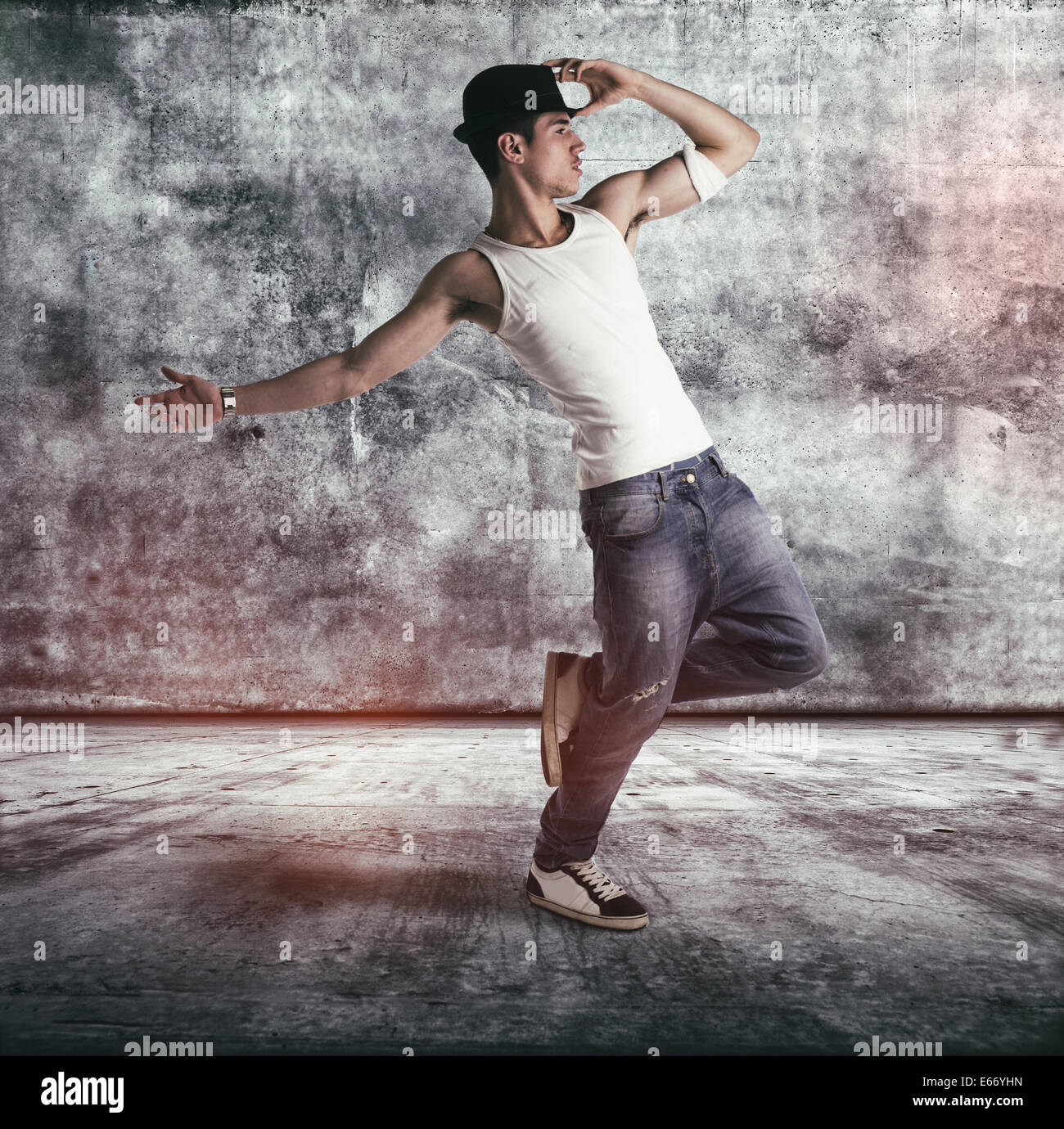 Hip young man in a tank top and hat doing a dance routine posing on one leg in a grunge concrete room or stage, profile full Stock Photo