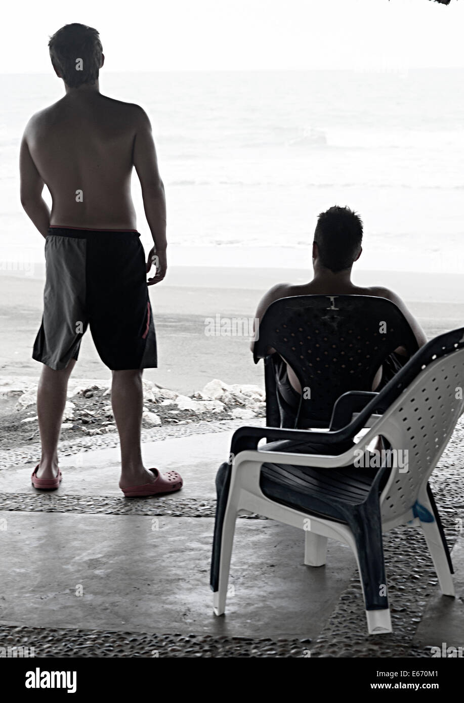 Two young men, twenties, watch the ocean and the waves covering the sandy beach in the early evening.  Cartagena, Colombia. Stock Photo