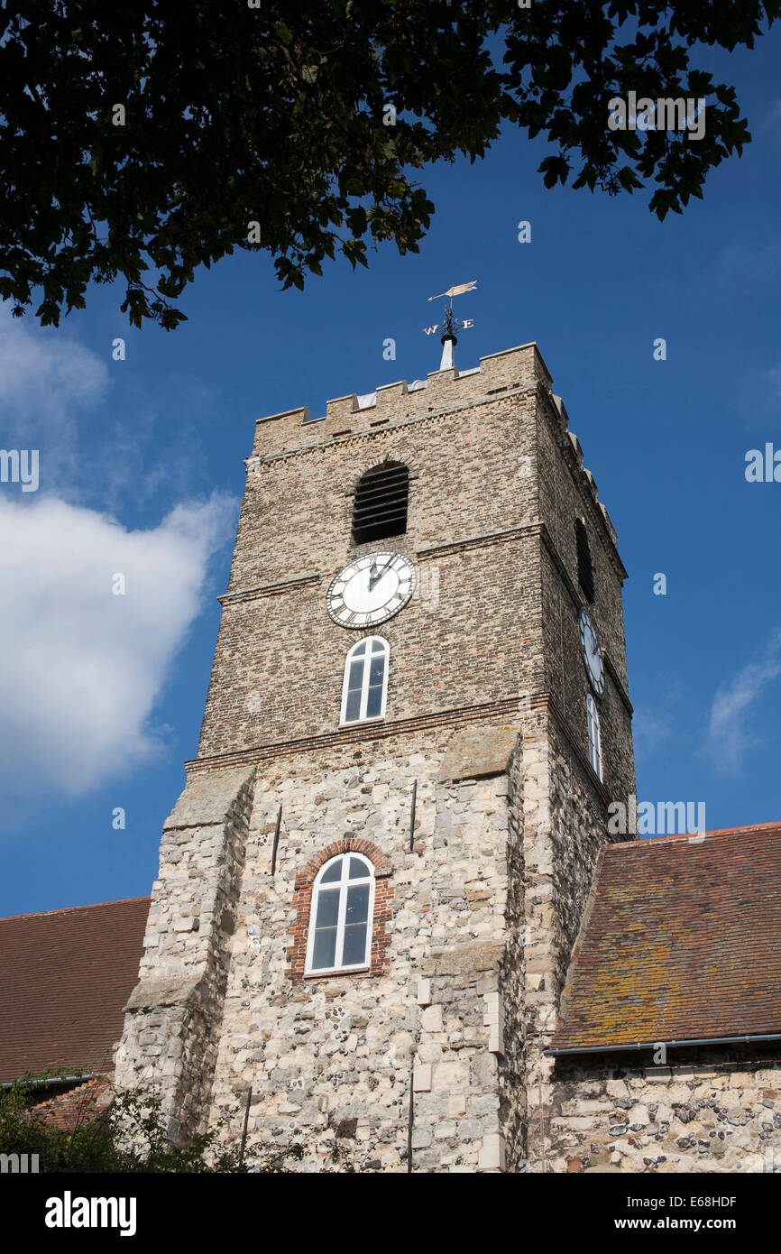 Detail of stone church showing tile roof, tower and clock on a sunny day with blue sky and clouds, with overhanging foliage Stock Photo