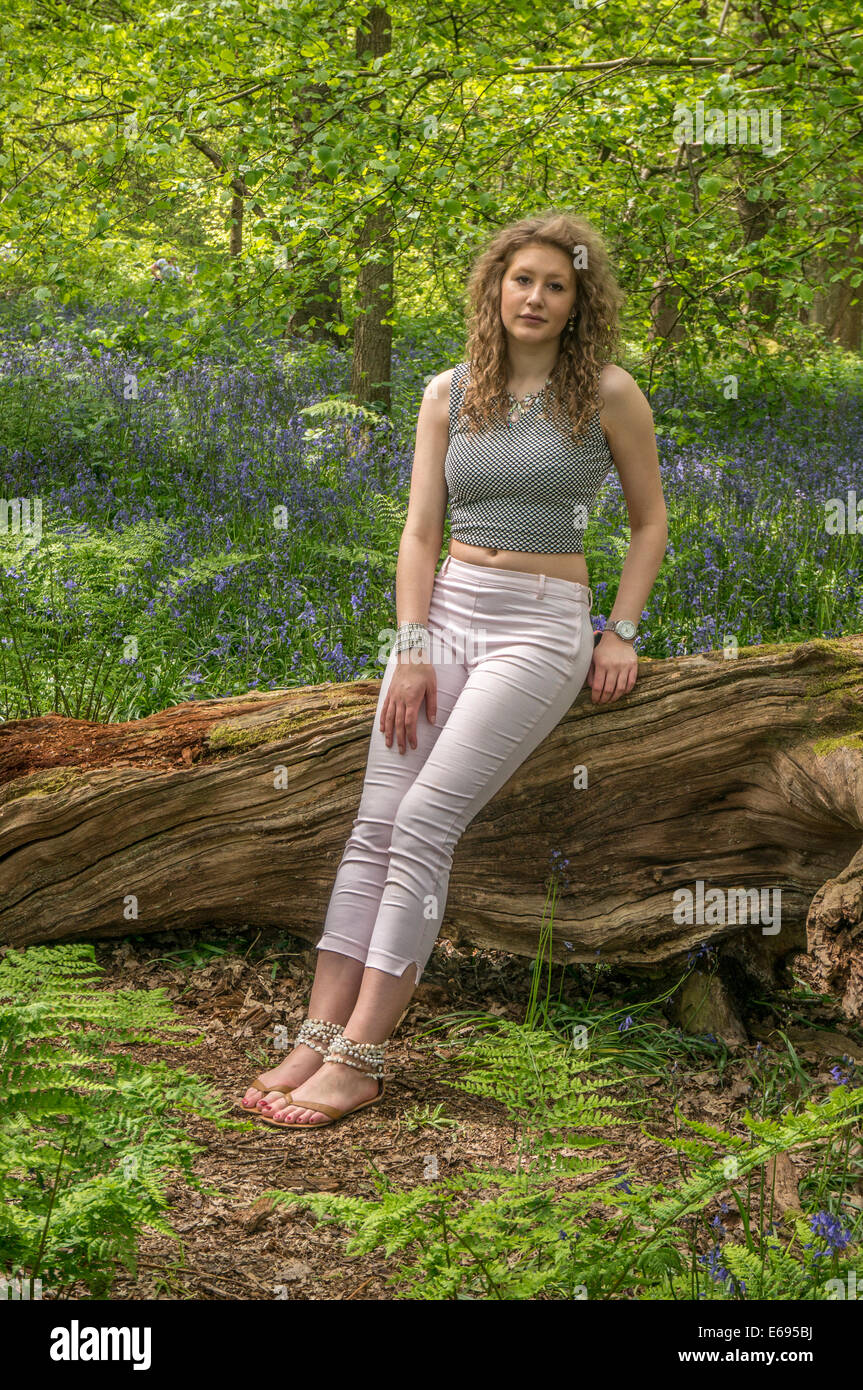 (Facing camera) A pretty teenage girl, with long, curly hair, sitting on an old tree amongst bluebells in the woods at Banstead, Surrey, England, UK. Stock Photo