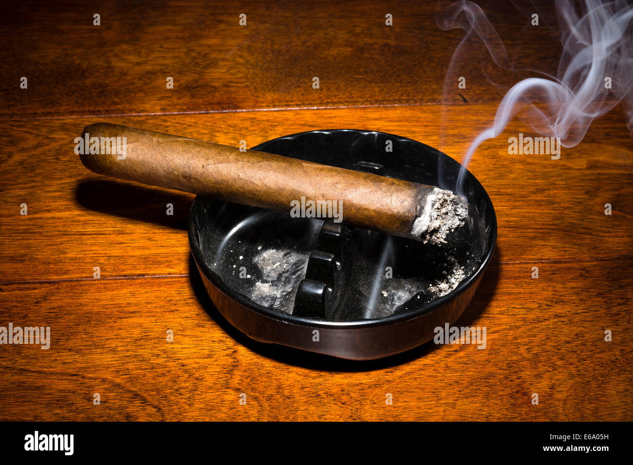 A burning cigar in a classic black ashtray streaming smoking in a dark, moody setting. Stock Photo