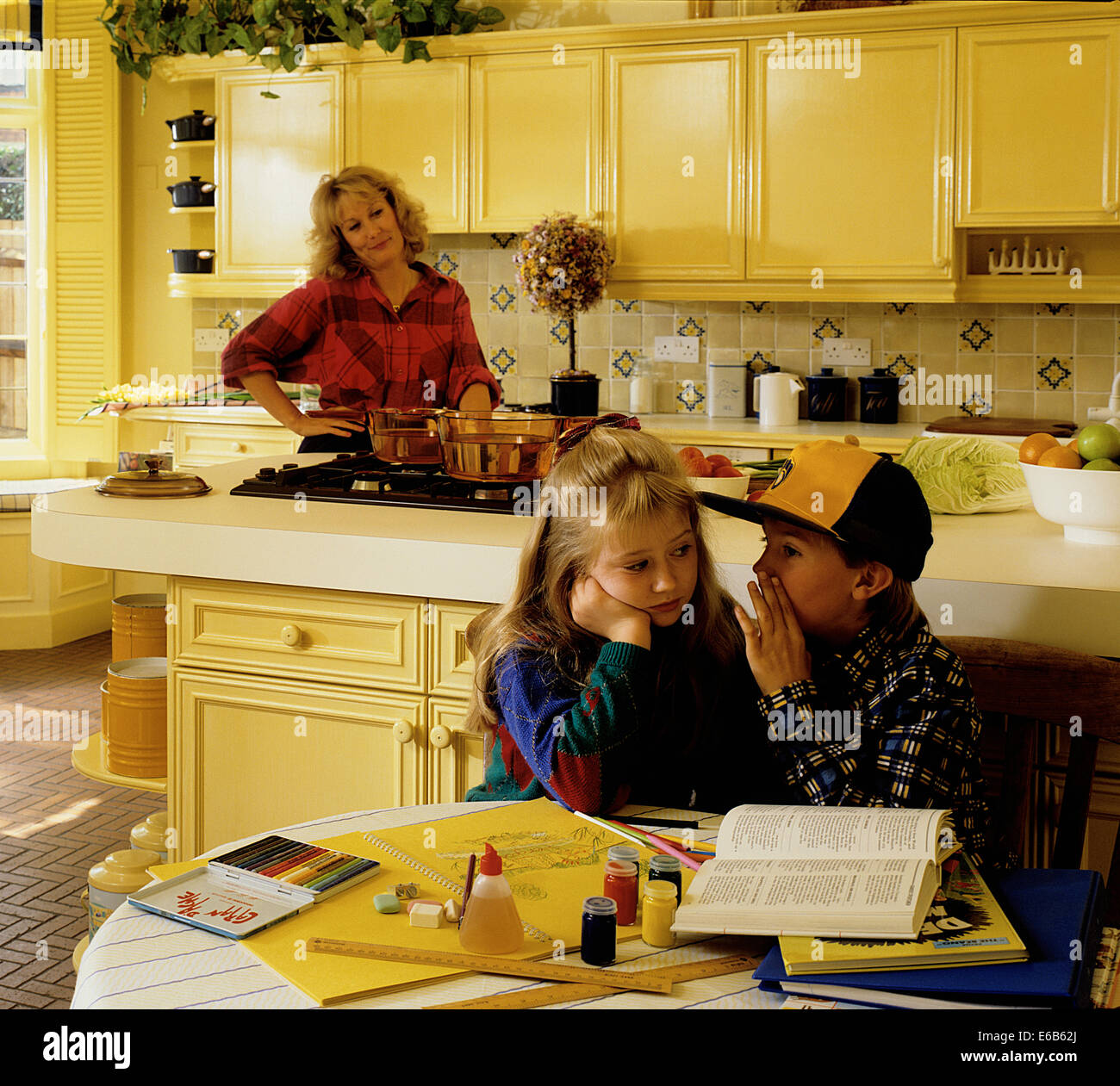 Young boy and girl sharing secret whilst mother looks suspiciously on, in modern home kitchen setting Stock Photo