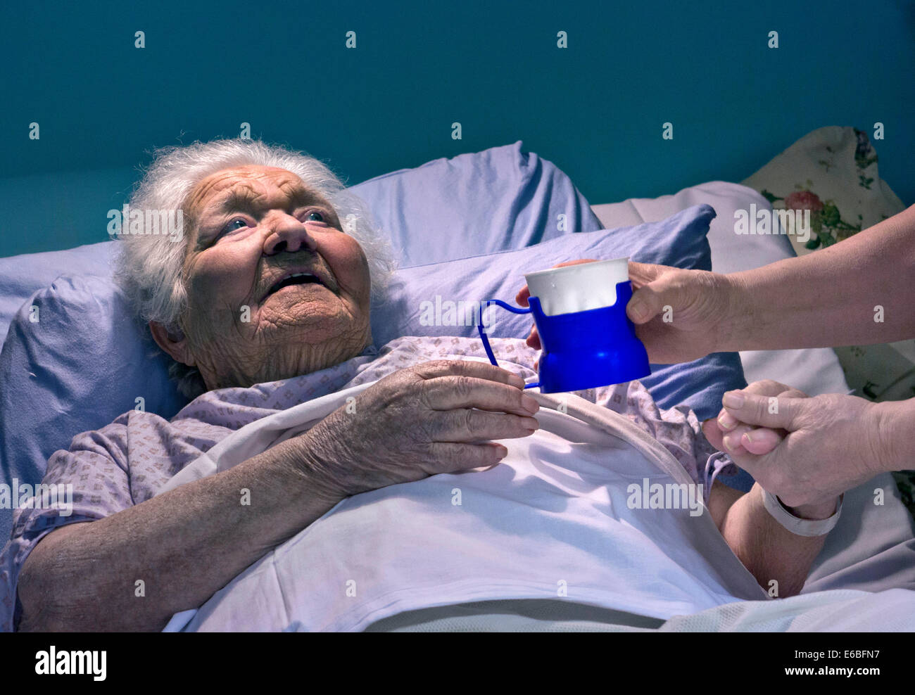 Contented smiling senior old age elderly lady in hospital bed comforting hand of carer nurse with hydration hydrating fresh cup of water Stock Photo