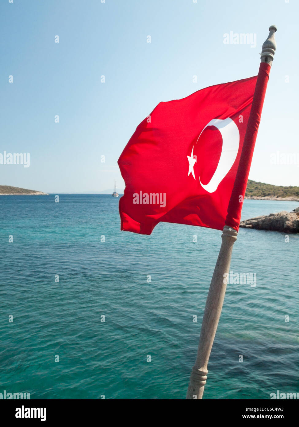 turkish flag on a hardwood mast flying in a strong wind with rocky coastline blue sea and boat behind Stock Photo
